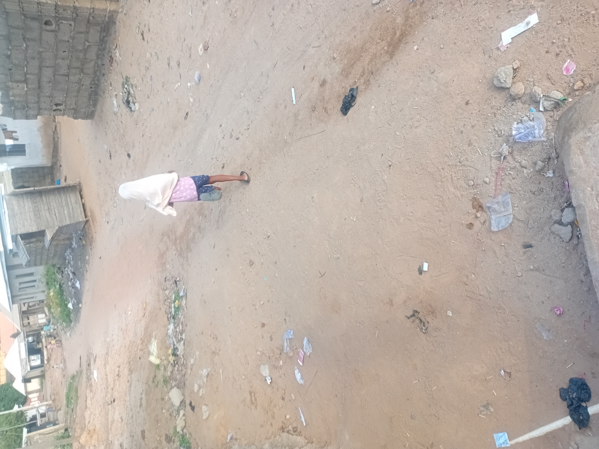 The image provides an overhead view of an unpaved, dusty ground in what appears to be a residential or semi-urban setting in Lokoja, Nigeria.
A single person is visible in the middle-left of the frame, seen from behind. This individual is wearing a light-colored, possibly white or off-white, head covering or garment that drapes over their upper body. They are also wearing patterned shorts, which appear to be purple or pink with white accents, and light-colored flip-flops or sandals. They seem to be standing or walking slowly away from the viewer.
The ground is composed of light brown soil or sand, indicating an unpaved road or open area. Scattered across this ground are numerous pieces of trash and debris, including various plastic bags (some black, some clear), small bits of paper or cardboard, and general refuse, suggesting an untidy environment.
On the left side of the image, several buildings are visible. These structures appear to be constructed from concrete blocks, with some showing wooden components. One building features a visible window. The architectural style and materials are consistent with informal or developing urban areas.
The lighting indicates it is daytime, possibly late morning or afternoon, with soft shadows. No specific activity or event is taking place beyond the person's presence in the scene. There is no legible text visible anywhere in the image.