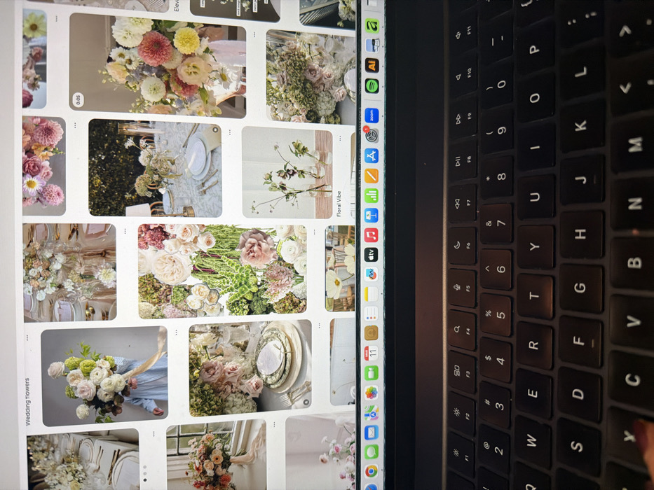 The image displays a close-up, top-down perspective of a dark-colored laptop, focusing on its backlit QWERTY keyboard and screen, in an active state. The scene is located in New York, United States of America.
The lower half of the frame is dominated by the laptop's black keyboard, featuring white character legends. A single finger, adorned with dark red nail polish, is partially visible at the bottom-left, resting near the 'Caps Lock' or 'Shift' key.
The laptop screen, visible in the upper half, displays a web browser, likely Safari, open to a Pinterest board titled "Wedding flowers." The board presents a grid of various image pins. These pins predominantly showcase floral arrangements, including large bouquets, elegant table settings with centerpieces, vases with minimalist flower designs, and collages of different flower types and greenery in an array of pastel and natural colors.
The macOS menu bar is present at the top of the screen, showing system icons such as Calendar (displaying "Mar 11") and a date indicator of "2023-11-05" further to the right. The macOS Dock is visible at the bottom of the screen, featuring application icons for services like Apple TV, Music, App Store, Zoom, Spotify, Adobe Illustrator, and Amazon, with some icons displaying red notification badges. The visible elements indicate a user actively browsing or researching floral and event design concepts.