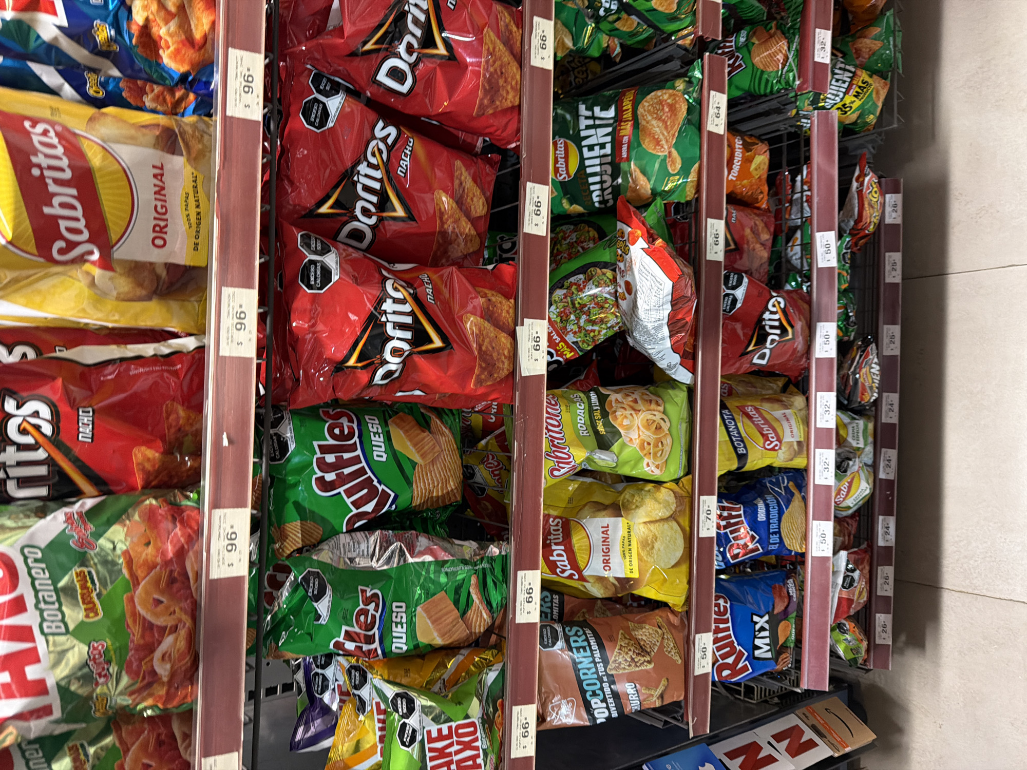 The image captures a close-up view of a snack food aisle, presumably in a store located in San Andrés Cholula, Mexico, as indicated by the prices in Mexican pesos on the shelf tags. The shelves are densely packed with various brands and flavors of chips. Brands such as Sabritas, Doritos, and Ruffles are prominently displayed. The shelves are metal wire racks with vertical maroon-colored price-tag holders along the front edge. The price tags indicate the cost of each item, with prices ranging from $24 to $96. The chips are packaged in colorful, sealed bags. The lighting appears to be standard store lighting. The overall setting is a retail environment.