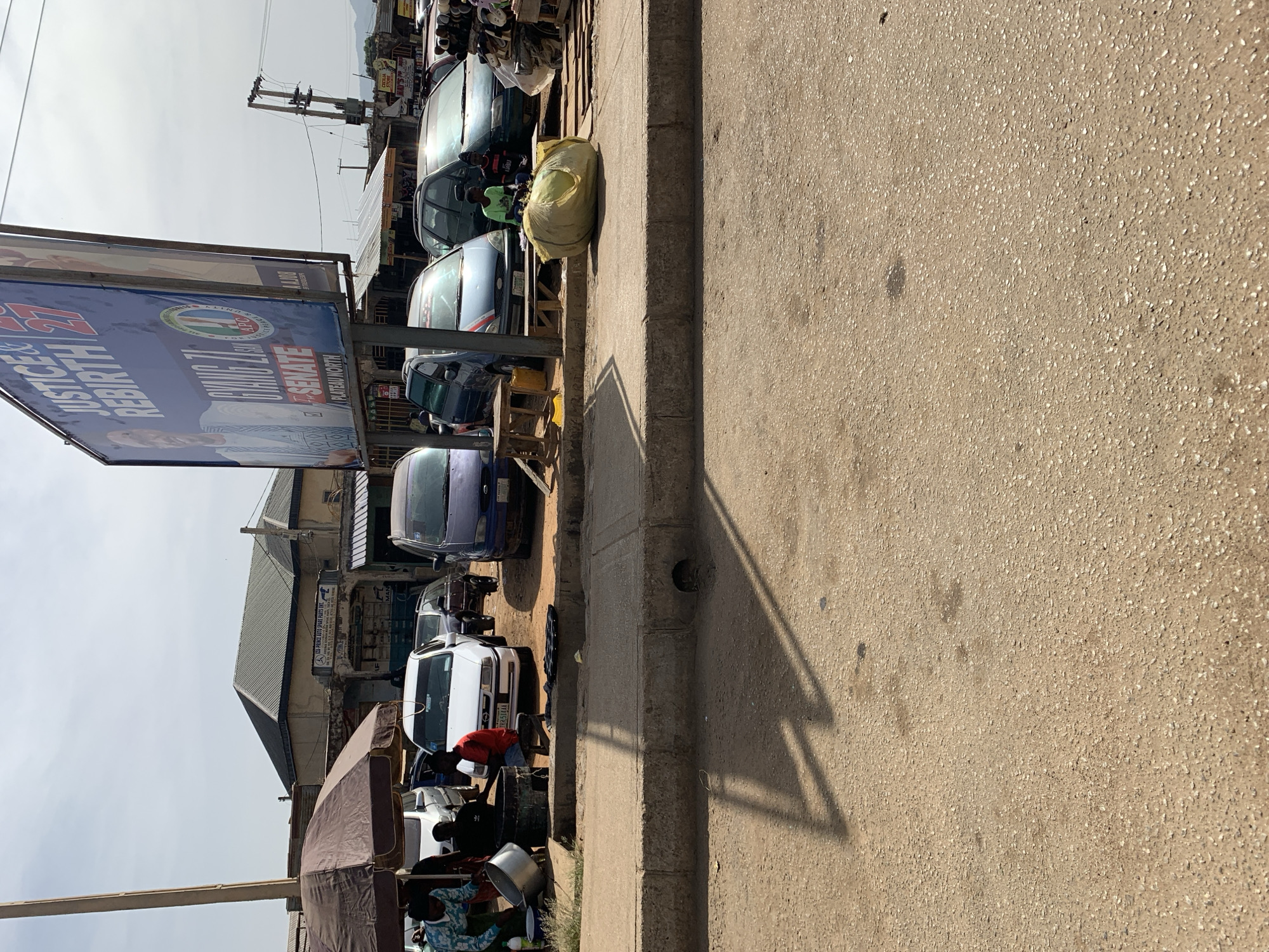 Here's a detailed description of the image:
The image depicts a street scene in what appears to be an urban area of Rafin Par, Nigeria. A row of parked cars lines the left side of the frame, next to a sidewalk. The sidewalk is composed of weathered concrete.
Several people are gathered around what appears to be a roadside food stall or small market operating under a brown tarp. One person is wearing a red shirt, and another is wearing a blue patterned shirt.
A large political billboard bearing the name "JUSTICE & REBIRTH" and "SENATE" is visible. The billboard is mounted on a metal frame.
Several parked cars are visible behind the food stall, including cars of white, blue, and green colors. A yellow bag sits on top of a wooden structure between two parked cars.
The storefront of "ZU-PRINCE AUTO SPARE PARTS ENTE" is visible in the background.
The street itself is asphalt. A shadow crosses the right side of the image. The sky is overcast. Power lines extend overhead.