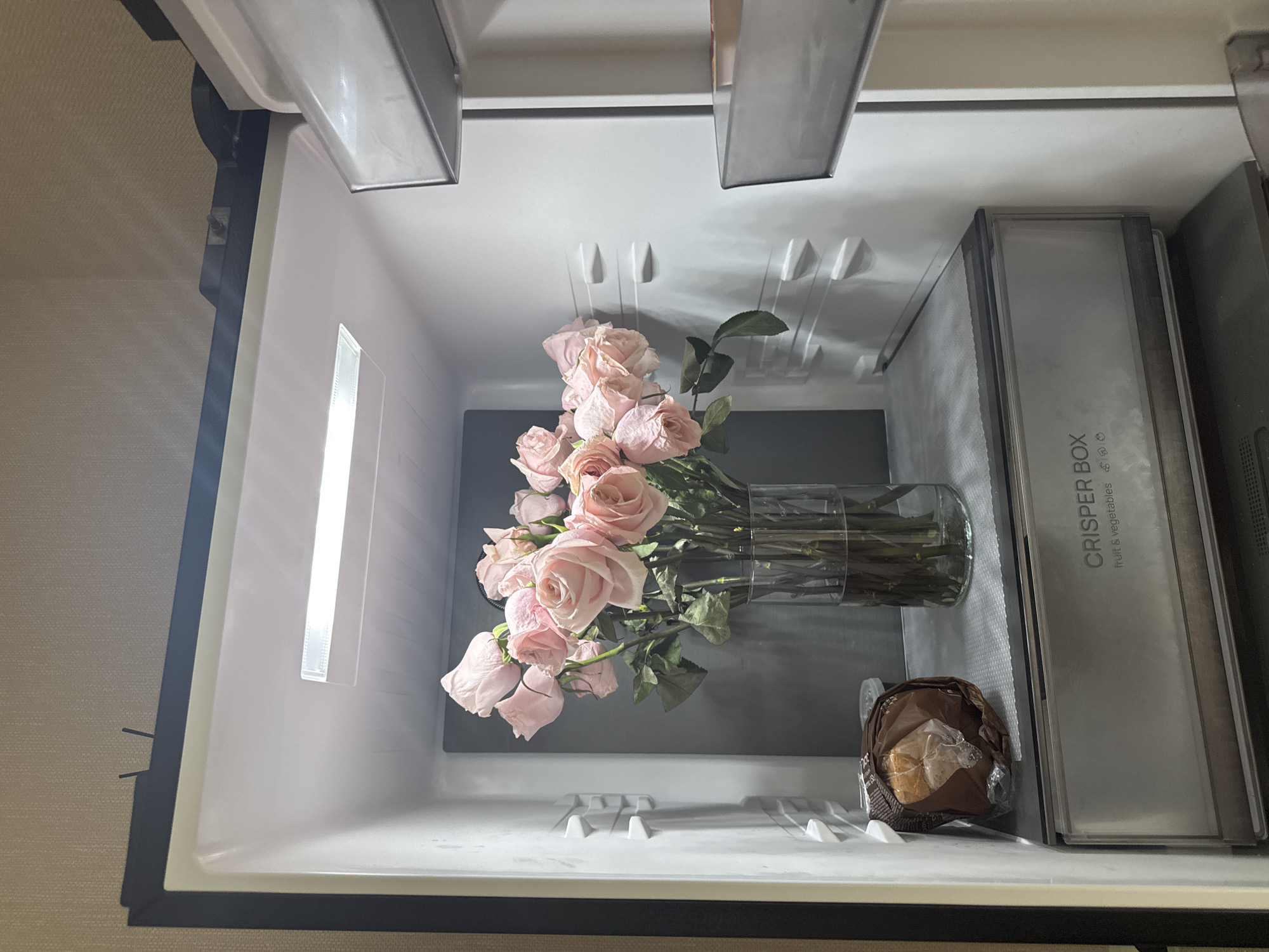 A bouquet of light pink roses in a glass vase sits on a shelf inside a refrigerator. To the left of the vase, a bread roll wrapped in brown paper packaging is visible. The refrigerator has a clear crisper drawer labeled "CRISPER BOX fruit & vegetables" at the bottom. The interior of the refrigerator is lit by an overhead light. This scene does not provide any information about the city of Perm, Russia.