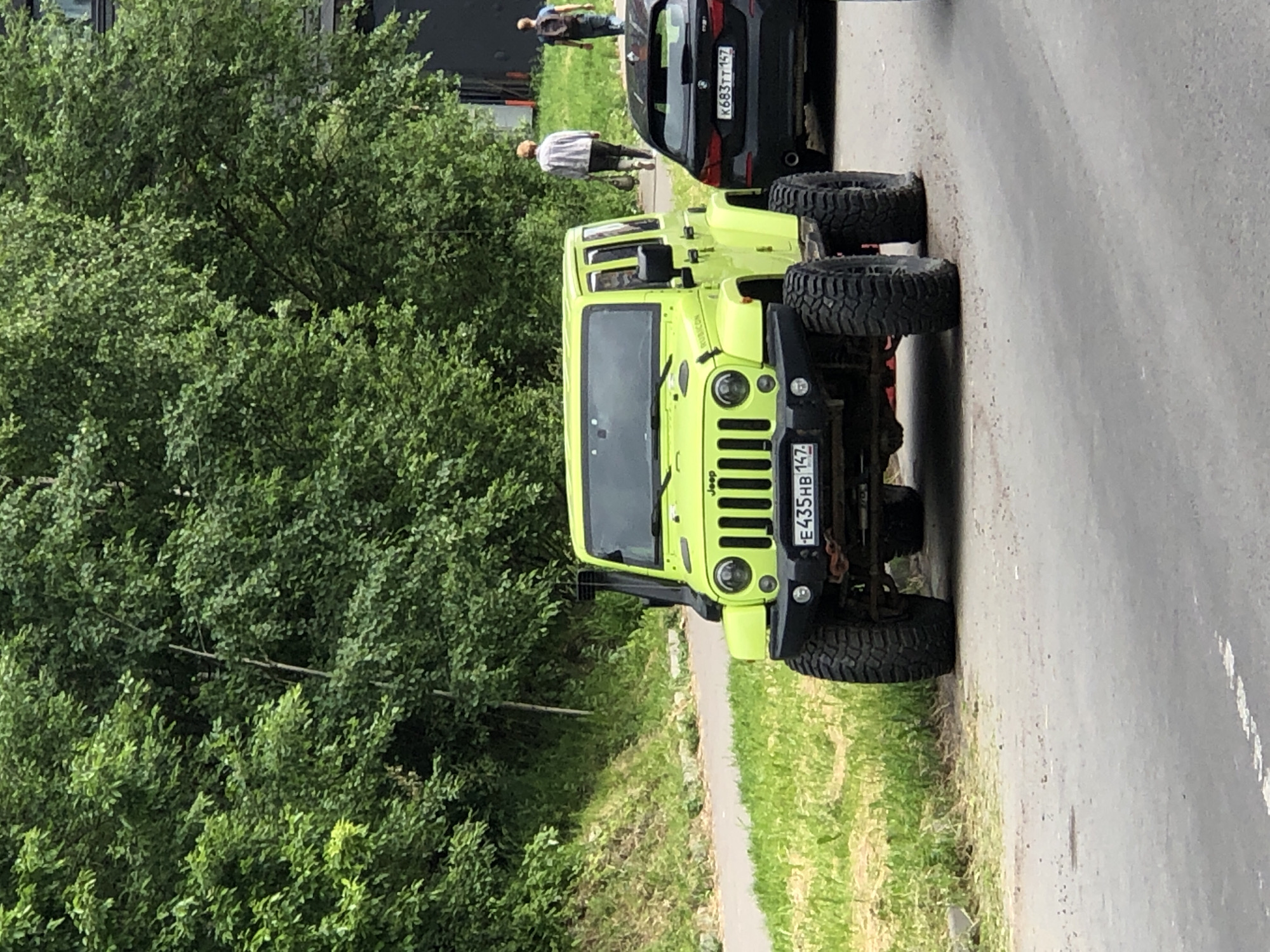A neon green Jeep Wrangler is parked on an asphalt road, facing forward. The Jeep has large, rugged tires and a snorkel on the driver's side. A license plate on the front of the Jeep reads "E435HB 147". To the right of the Jeep, a black BMW sedan is parked. In the background, two people are walking away from the camera on a sidewalk. Behind the sidewalk is a lush green area with trees and some buildings. The scene appears to be outdoors, possibly in a park or a suburban area.