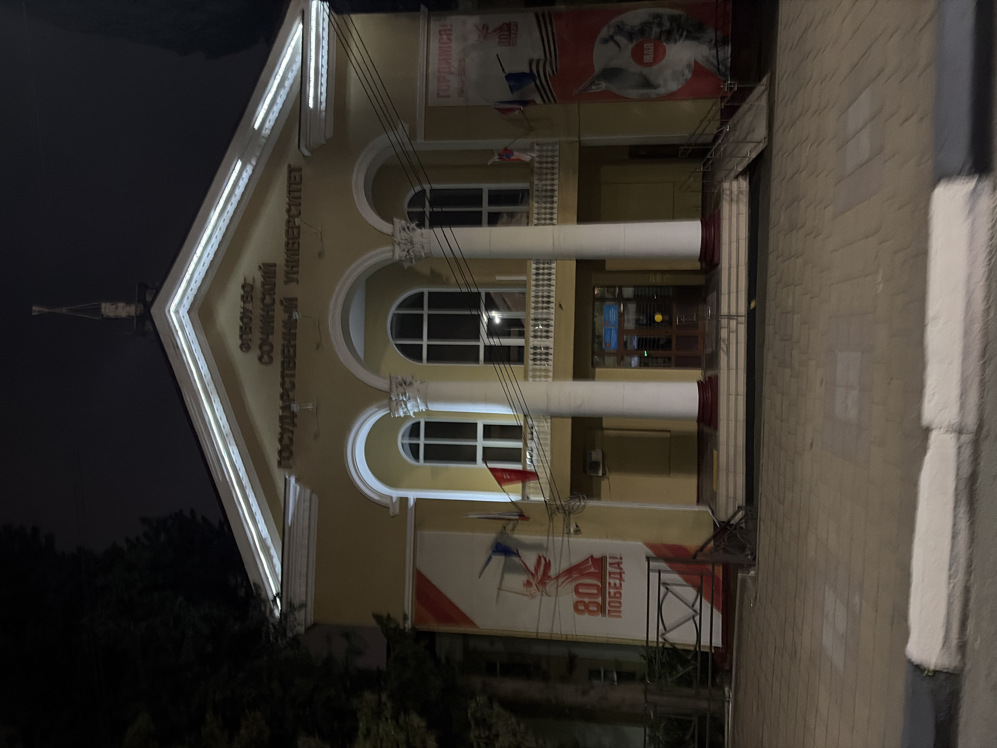 The image shows the facade of a yellow building at night, likely a public institution or university. Above the entrance, there is signage that reads "СОЧИНСКИЙ ГОСУДАРСТВЕННЫЙ УНИВЕРСИТЕТ" (Sochi State University). The building features a central portico with three arched windows above, flanked by two large white columns. To the left of the entrance, a large banner displays the number "80" and the word "ПОБЕДА!" (Victory!), indicating a commemoration of an 80th anniversary of victory. Two flags, one red and one with the Russian flag, are mounted on the facade. To the right, another banner displays the word "ГОРДИМСЯ!" (We are proud!) and a circular image. A ramp leads up to the entrance, suggesting accessibility for individuals with disabilities. The sidewalk in front of the building is paved with interlocking stones. A dark tree is visible to the left of the building. The overall lighting suggests it is nighttime, with artificial lights illuminating the building.