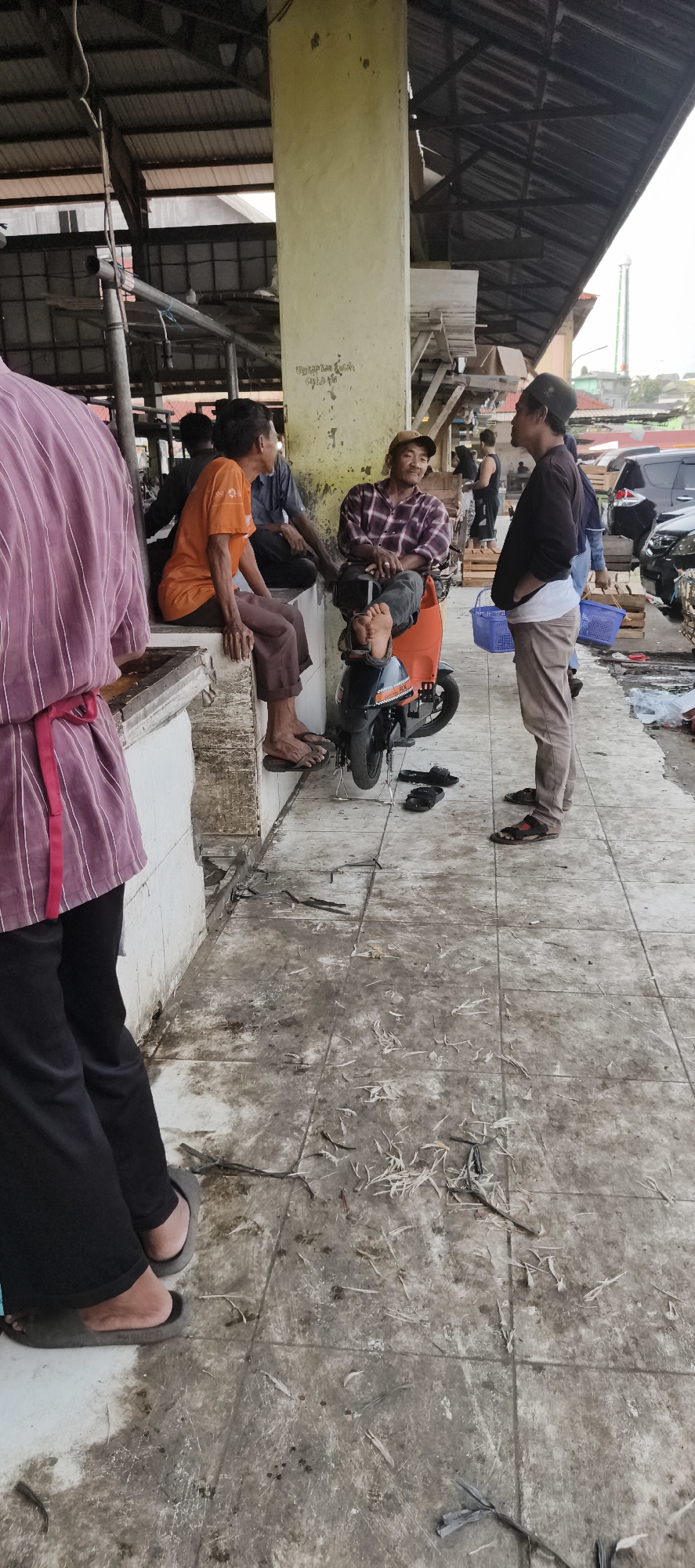 The image depicts a scene likely within a bustling market in Jambi City, Indonesia. Four men are visible, with three engaged in conversation and one standing apart. One man, wearing an orange t-shirt and brown pants, is seated on a raised concrete platform. Another man in a plaid shirt and jeans is seated on an orange electric scooter, his feet resting on the ground. A third man, wearing a black cap and brown pants, stands and talks with the man on the scooter. In the foreground, a person in a purple striped shirt and black pants is partially visible. The environment suggests an outdoor or semi-covered market setting, with a tiled floor that appears somewhat dirty and littered with debris, possibly from fish or produce. Wooden crates and blue baskets are present in the background, along with parked vehicles. The overall impression is of a casual gathering amidst a commercial area.
