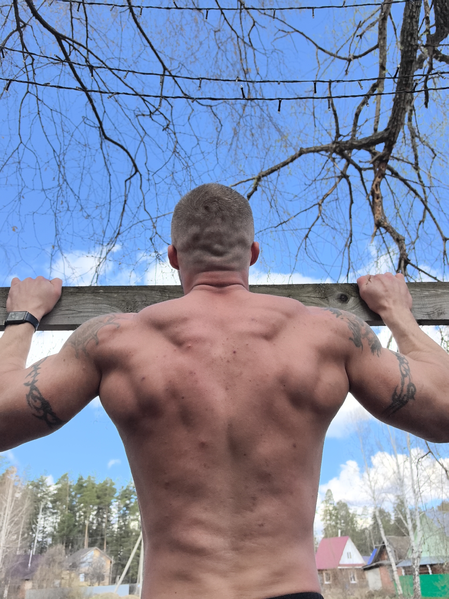 This image, set in Noviy, Russia, captures a shirtless man from behind, engaged in an outdoor physical activity. He is holding onto a rough-hewn wooden plank with both hands, positioned as if performing a pull-up or similar exercise. The man displays a muscular back and shoulders, with prominent tattoos on both upper arms. A dark-strapped smartwatch is visible on his left wrist. His hair is short and neatly cut.
Above the man, a bright blue sky is dotted with scattered white clouds. Bare tree branches, characteristic of late winter or early spring, stretch across the upper portion of the frame. String lights with small bulbs are interwoven among these branches, suggesting an outdoor lighting setup.
The background reveals a rural or suburban landscape under natural daylight. On the left, evergreen pine trees and parts of houses are visible. To the right, deciduous trees, including birches, are prominent, along with additional houses featuring distinct roofs (one red, one green). A fence and a utility pole are also discernable in the middle distance. The scene conveys an individual's engagement in outdoor fitness within a natural, rustic environment during daytime. No text is visible in the image.