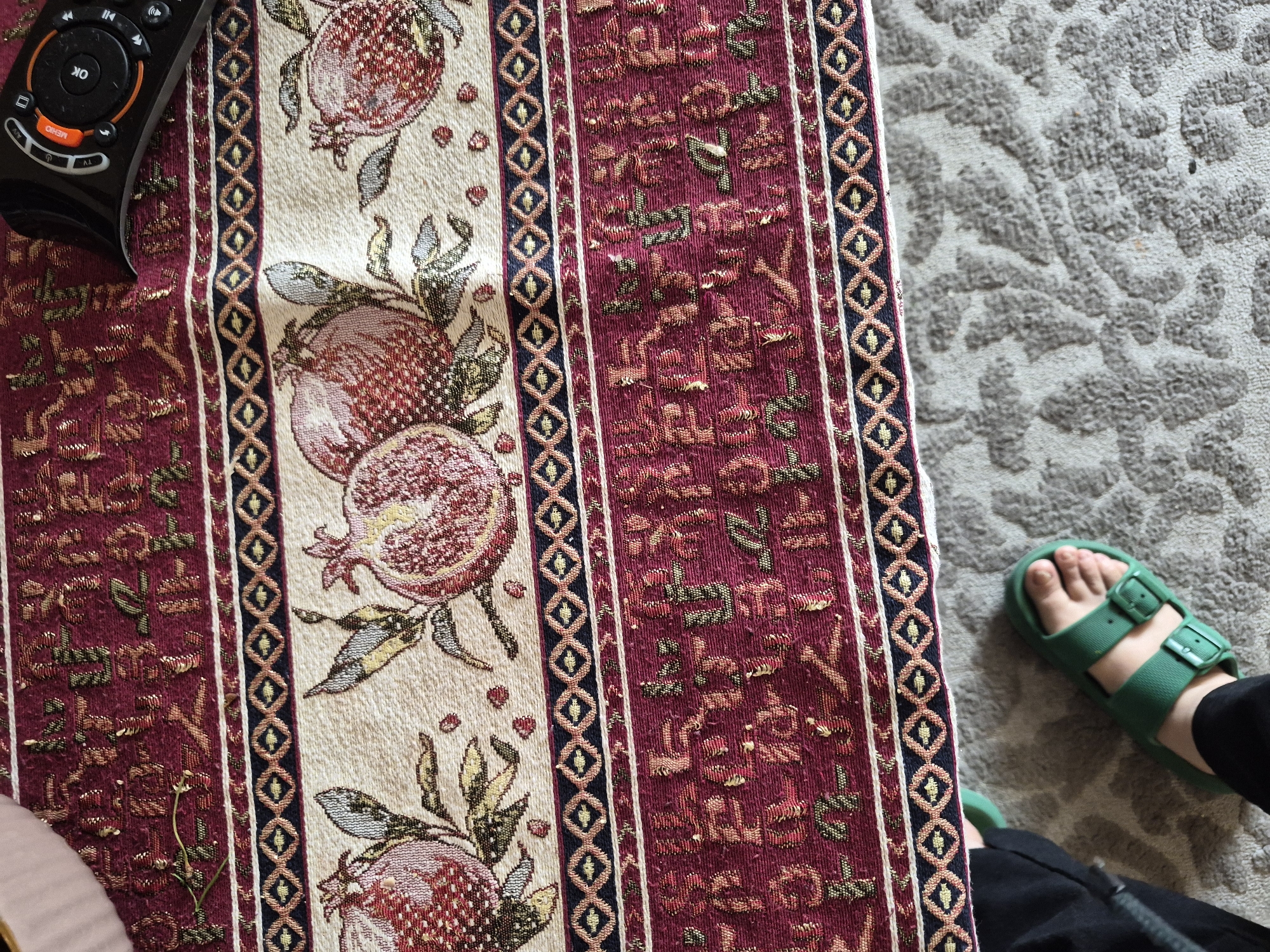 A top-down indoor photograph shows a person's foot, a TV remote, and an elaborately patterned fabric runner resting on a textured rug. The fabric runner features a central cream-colored stripe adorned with multiple red pomegranates and green leaves. This stripe is bordered on both sides by a narrow dark blue or black band containing a repeating gold diamond motif. Adjacent to these, on either side, are wider maroon-colored sections intricately woven with stylized symbols resembling ancient Egyptian hieroglyphs in gold, green, and red threads.
In the upper left corner, a black TV remote control is partially visible, resting on the fabric. The remote has prominent "OK" and "MENU" buttons in orange and white, surrounded by a directional pad. In the lower right of the frame, a person's foot, clad in a bright green two-strap sandal, rests on a light grey rug with a raised, organic, or abstract pattern. The overall setting suggests a relaxed indoor environment, possibly a living room, in Yerevan, Armenia. The lighting indicates an interior scene, with no specific time of day discernible.