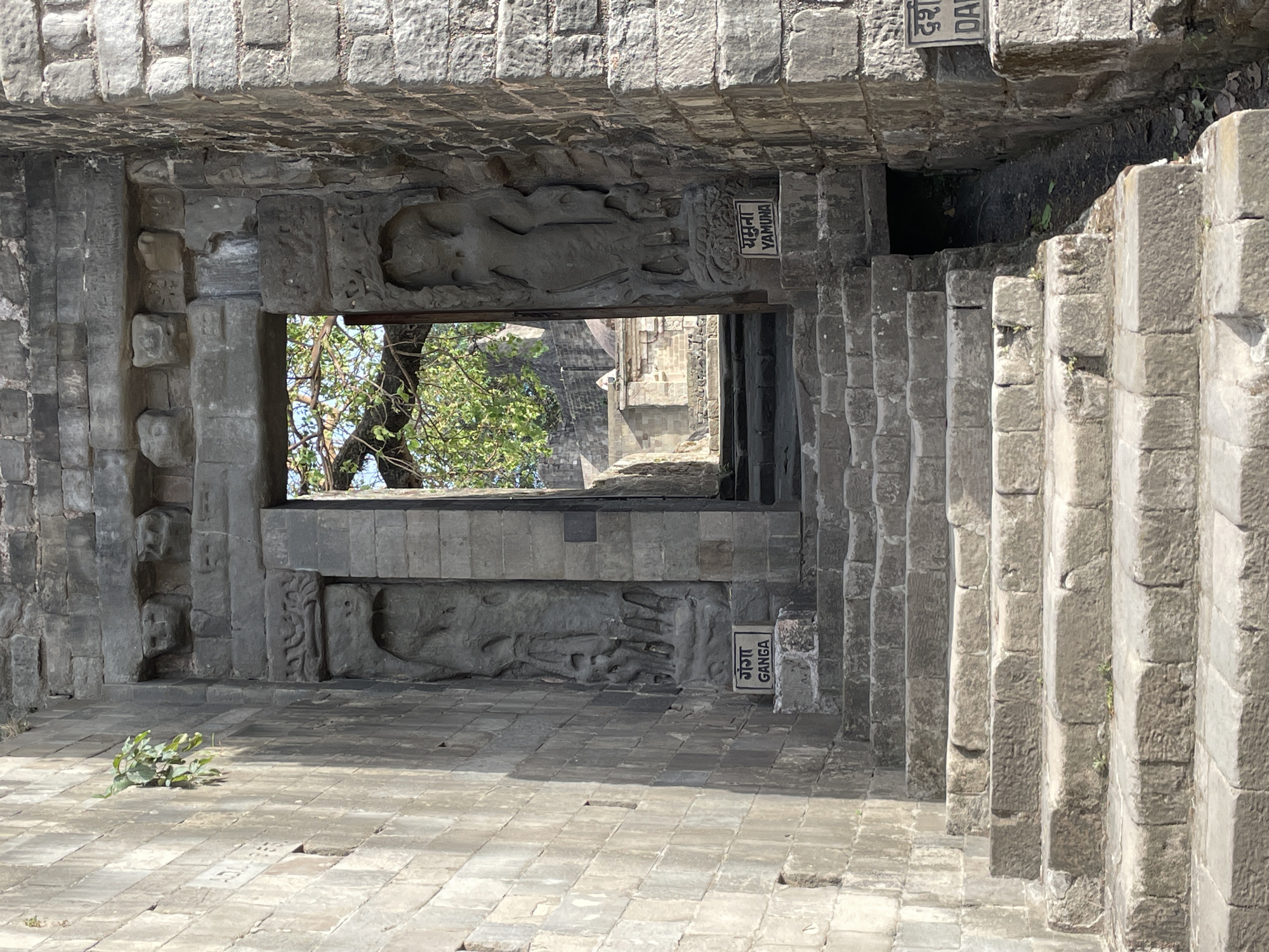 This image depicts a stone structure, possibly part of a cave or ancient temple, in Kaswara, India. The structure features carved figures of goddesses, identified as Yamuna and Ganga by adjacent signs, framing an opening that reveals a view of trees and a distant stone building. A series of steps ascends to the right of the structure. The ground is paved with stone tiles, and a small plant grows in the foreground. The scene appears to be illuminated by daylight.