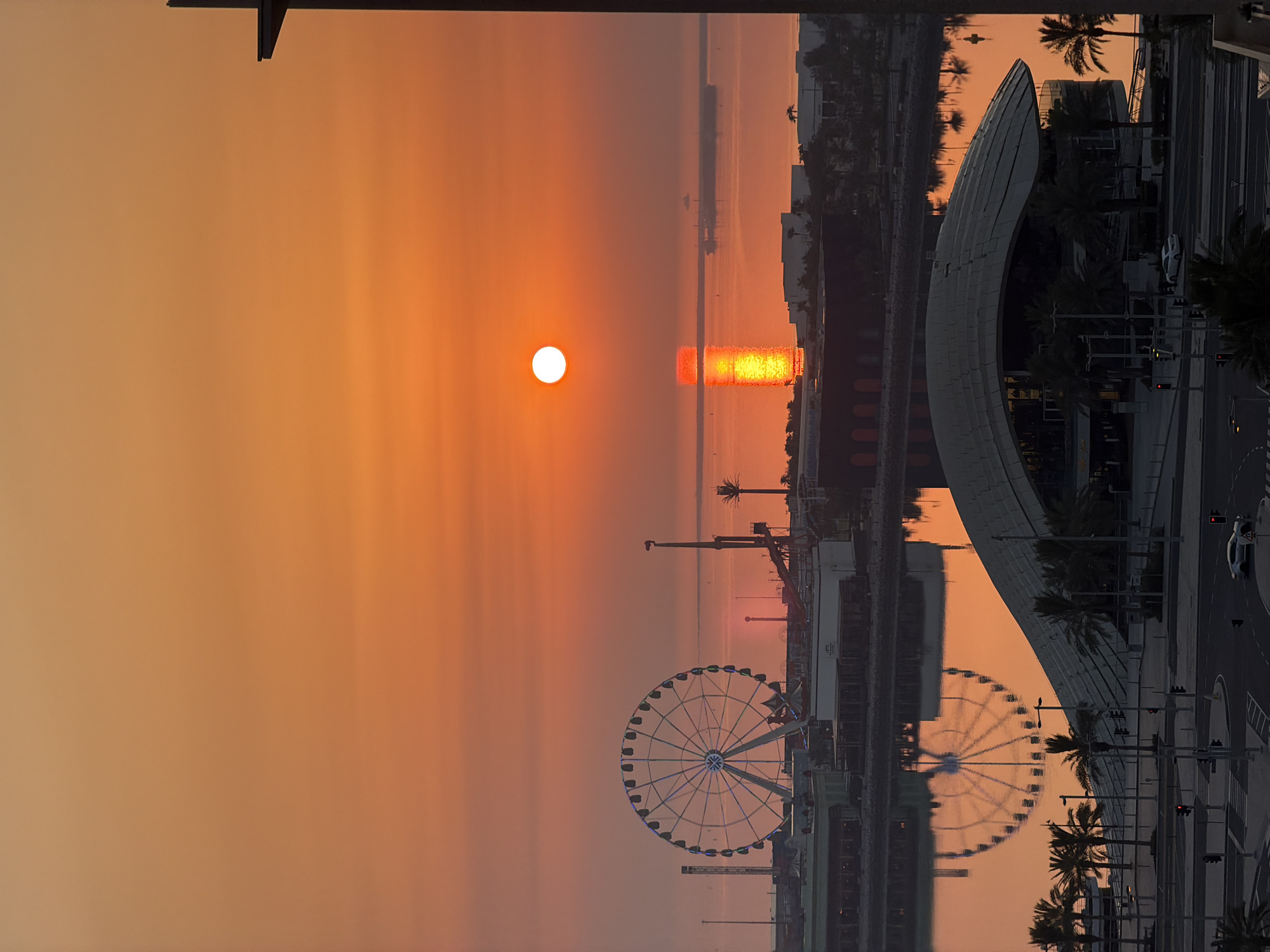 The image shows a cityscape at sunrise, with the sun casting a vibrant orange glow across the sky and reflecting on the water. In the foreground, a modern building with a curved roof is visible, flanked by palm trees. Beyond this, a body of water reflects the scene, including a Ferris wheel and various other structures. In the distance, a large ship is present on the water, and more buildings line the coast. The overall impression is one of a serene and picturesque urban sunrise over a waterfront area.