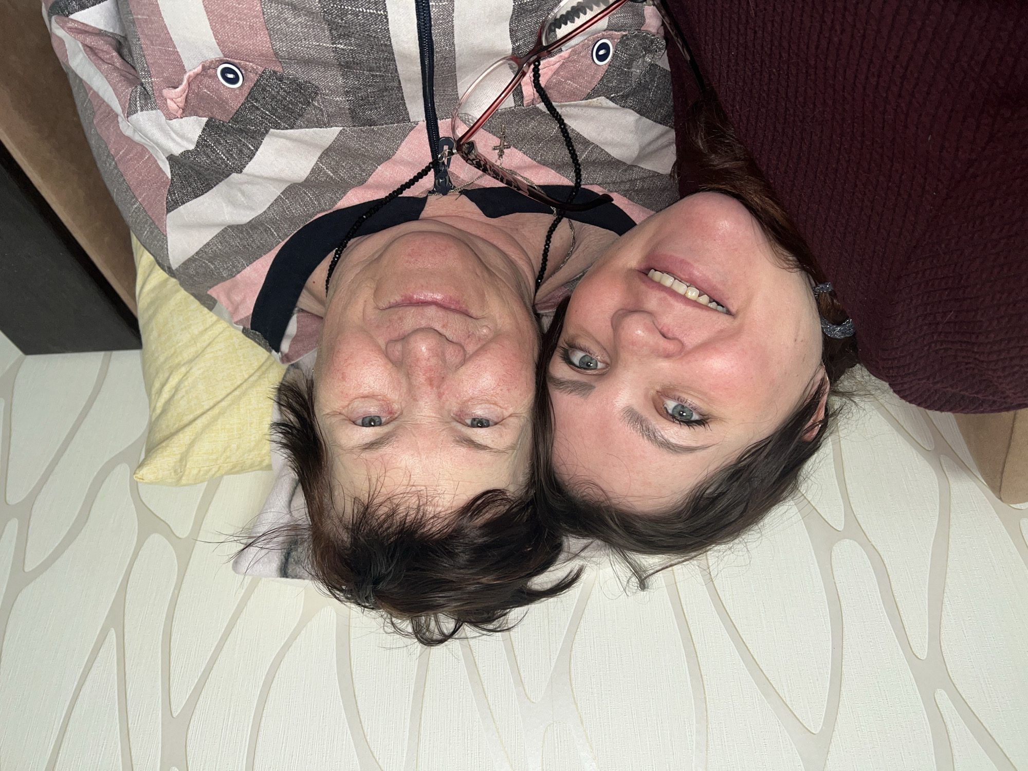 Two women are lying head-to-head on a surface with light-colored wallpaper featuring a branching pattern. The woman on the left wears a striped shirt with a zipper and glasses with a beaded necklace. The woman on the right wears a burgundy-colored shirt and is smiling. The setting might be indoors based on the wallpaper. There is no indication of Perm, Russia, or any city.