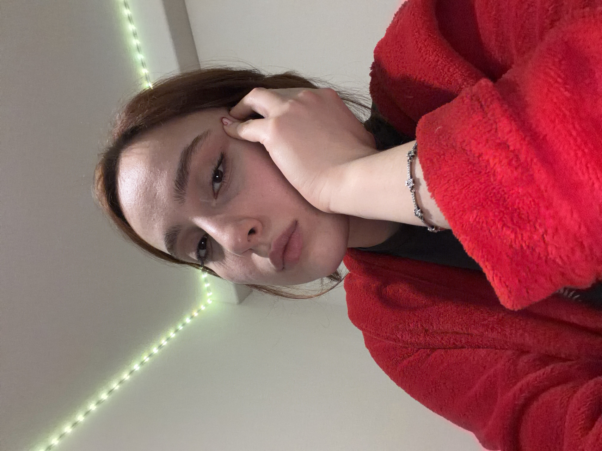 This image presents a close-up, slightly low-angle portrait of an individual in an indoor location within Strasbourg, France. The subject, with fair skin, brown hair, and dark eyes, faces forward, making direct eye contact with the viewer. Their right hand is positioned to support the right side of their face, with the fingers resting against the cheek and temple. A small pink heart design is visible on the thumbnail of the right index finger. The individual is wearing a vibrant red, textured garment, likely made of fleece, over a darker top. A silver-colored bracelet adorned with star-shaped links is present on the right wrist. The background features a plain, light-colored wall. A string of green LED lights is affixed to the corner where the wall meets the ceiling in the upper left quadrant of the frame. The scene is illuminated by soft, frontal lighting.