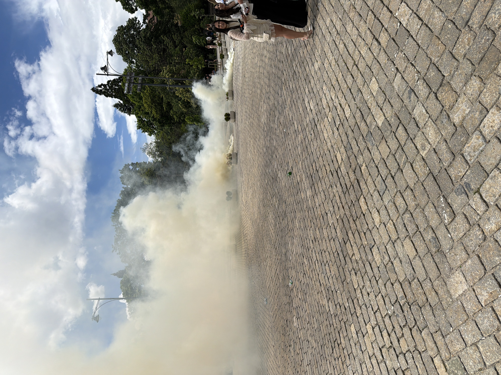 The image shows an outdoor scene, possibly a celebration or event, on a paved brick area in Sofia, Bulgaria. Two women are seen standing to the upper right corner of the image, partially obscured. The background features trees, a sky with clouds, and what seems to be a lamp post. A significant amount of yellowish smoke or fog fills the middle ground of the image, partially obscuring the view.
