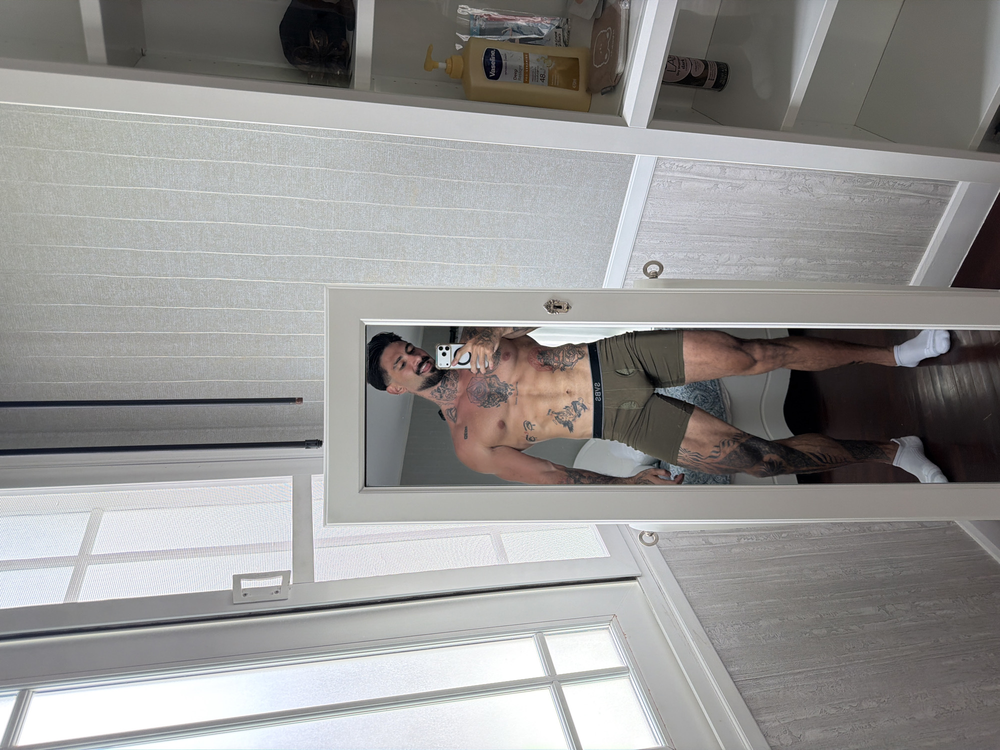 A tanned man is taking a mirror selfie indoors. He is wearing underwear and white socks. He has tattoos covering his torso and left leg. A phone can be seen in the mirror. The room has a white cabinet with toiletries. There is a window in the bottom left of the image. The location is in Chalong, Thailand.