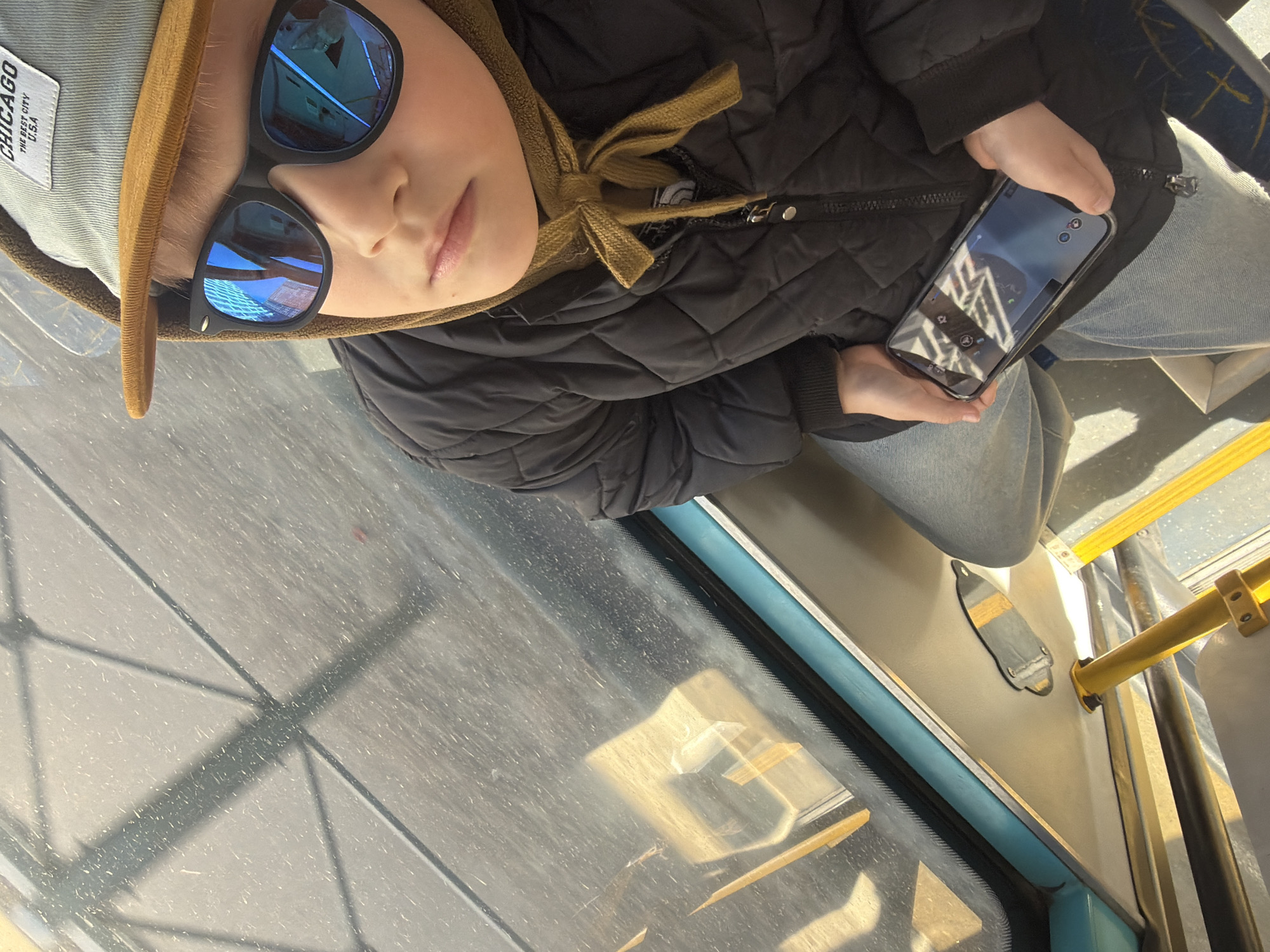 A child wearing a hat and sunglasses is sitting on public transportation, holding a phone. The phone screen reflects the interior of a building. The scene is illuminated by sunlight. The interior of the bus includes a window, poles, and seats. The location is Saint Petersburg, Russia, as evidenced by buildings reflected in the glasses and phone screen.