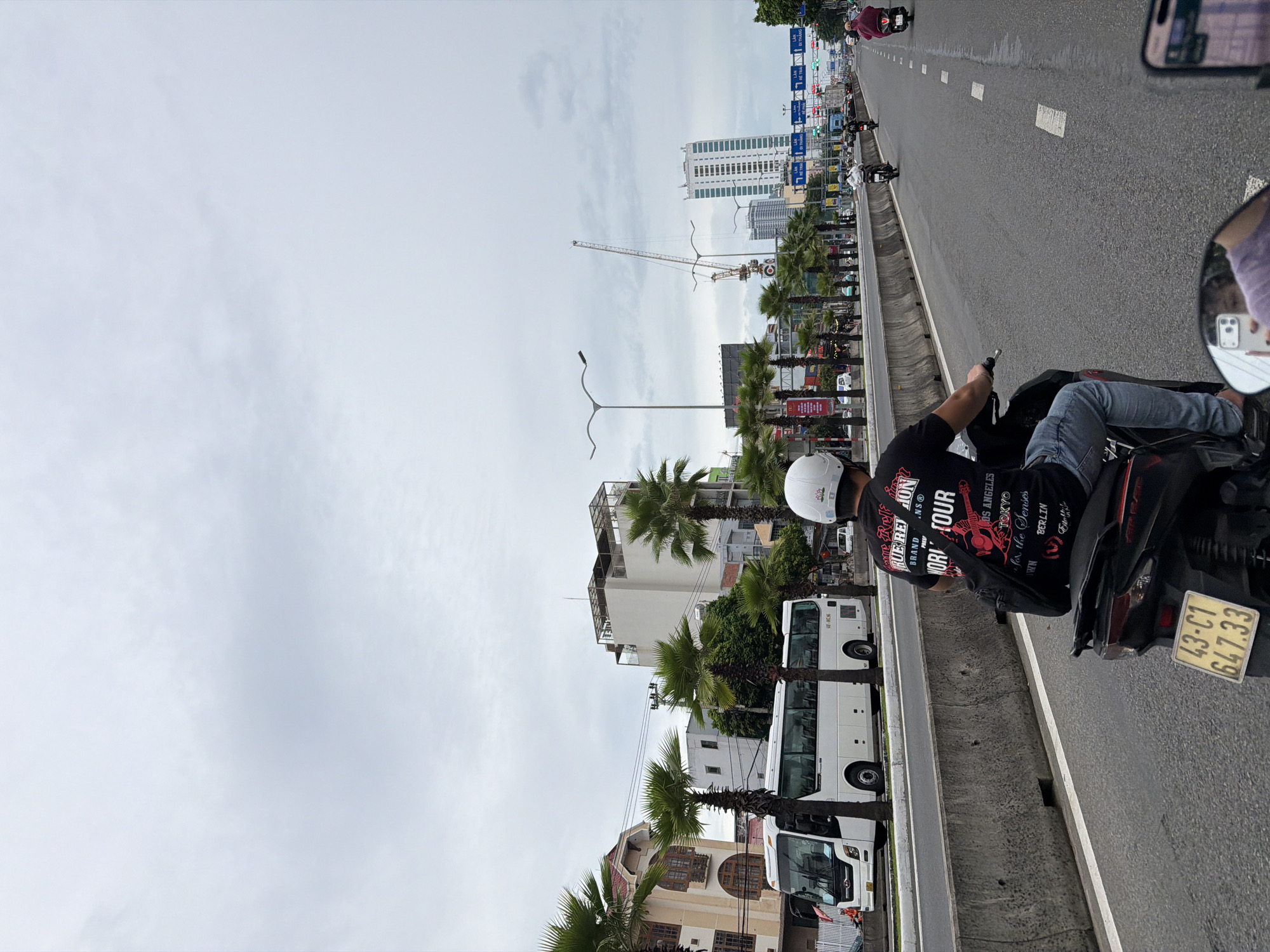 The image depicts a street scene in Da Nang, Vietnam, likely during an overcast day. A person on a motorcycle, wearing a white helmet and a black t-shirt with red lettering that reads "TRUE REFLECTION WORLD TOUR," is the main subject in the foreground, facing away from the camera. Their blue jeans and the motorcycle's license plate, "43-C1 647.33," are clearly visible. The motorcycle is in motion on a paved road, with other vehicles, including another motorcycle and a bus, in the background. Palm trees line the street, and several buildings, including a tall, modern structure and a crane in the distance, suggest an urban environment. Traffic lights and street signs are visible, indicating an active thoroughfare. The overall atmosphere is that of a typical day in a bustling Vietnamese city.
