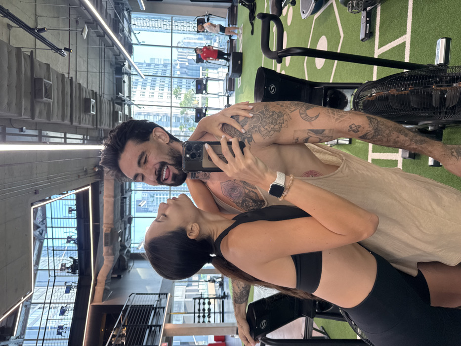 This image captures a mirror selfie of a female and a male in a modern high-rise gym facility in Bangkok, Thailand.
The female, on the left, has dark hair in a ponytail and is dressed in a black sports bra and black athletic shorts. She is leaning into the male, with her face near his neck in an affectionate posture. Her right arm is wrapped around his upper back. Her left hand holds a dark-colored smartphone, equipped with a clear case, to take the photograph. She wears a white smartwatch and a beaded bracelet on her left wrist.
The male, positioned on the right, has dark hair, a beard, and a mustache. He is wearing a light-colored sleeveless tank top and displays numerous tattoos across his arms, chest, and neck. He is looking towards the phone camera with a playful expression, mouth open, and tongue slightly extended. His left arm is around the female's lower back.
The gym features a green artificial turf floor with white markings. Visible equipment includes an air bike in the foreground, and multiple treadmills positioned near large, floor-to-ceiling windows in the background. These windows offer a clear view of a dense cityscape composed of modern high-rise buildings under a bright sky. The ceiling displays exposed concrete, ventilation ducts, and track lighting. In the background, other gym users are present, including one individual in a red shirt near a treadmill and another further right.