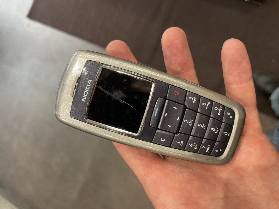 A person is holding an old, silver-colored Nokia mobile phone in their right hand. The phone is positioned diagonally across the palm, with fingers curled to support it from underneath and the thumb resting on the right side. The background is a blurry, warm brown and grey, suggesting an indoor setting with perhaps a floor or carpet.
The Nokia phone itself appears well-used and shows signs of wear. The front panel, which is a darker grey/black, features the "NOKIA" logo prominently above the display screen. The screen is dark, indicating it is off or unpowered, and is noticeably cracked with several prominent lines radiating across its surface. There are also numerous scratches and smudges on the screen and the surrounding bezel.
Below the screen is a standard alphanumeric keypad. Keypad buttons are black with white lettering. Visible keys include a "C" button, a green button with a phone icon (likely for making calls), and a red button marked with a circle and a vertical line (typically for ending calls or power). The number keys from "0" to "9" are present, along with the asterisk "*" and hash "#" keys. Each number key also displays corresponding letters for text input: "2 abc", "3 def", "4 ghi", "5 jkl", "6 mno", "7 pqrs", "8 tuv", "9 wxyz". The phone's casing is chipped and scratched, particularly around the edges and near the screen, further emphasizing its age and extensive use.