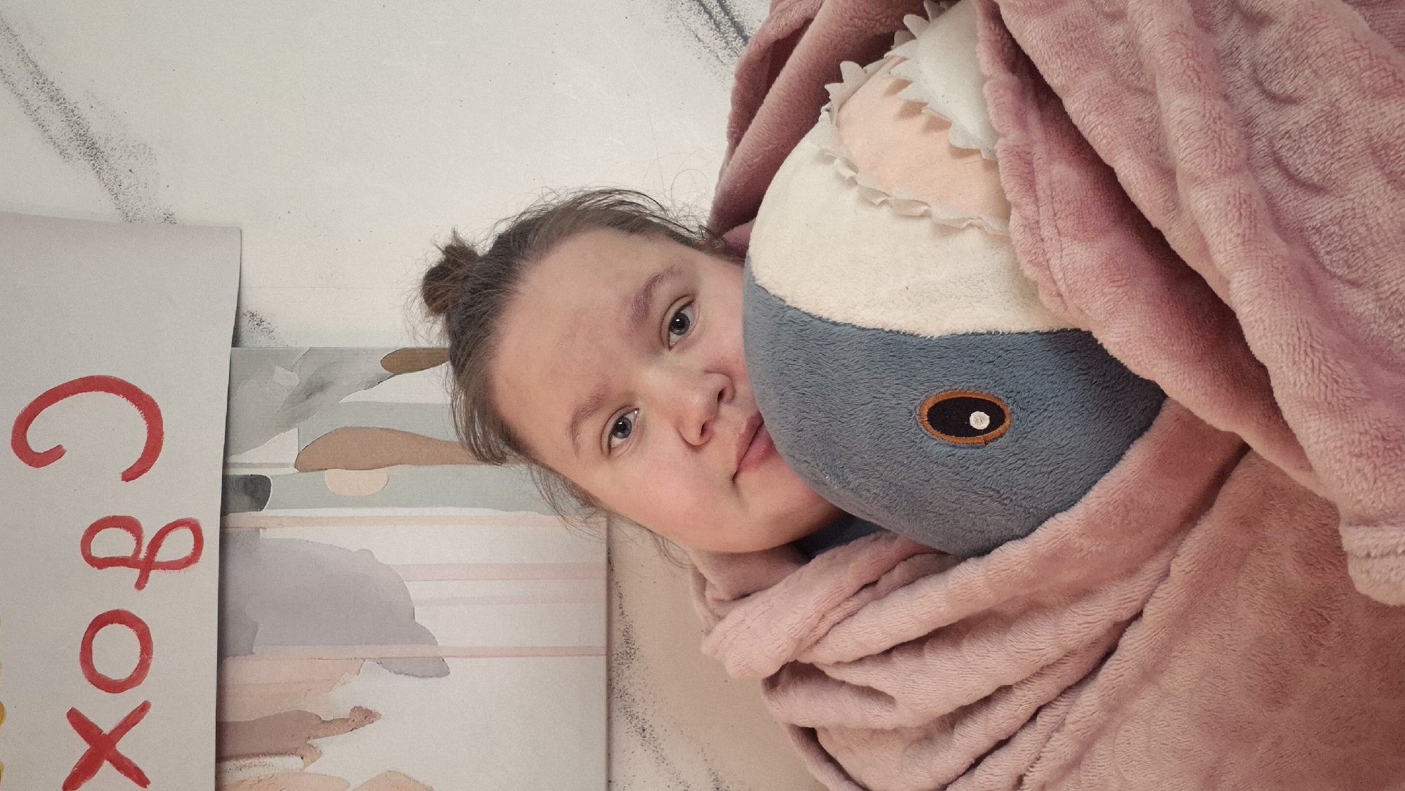 A girl with her hair in a bun, wearing a pink robe, is holding a stuffed shark toy. Next to her is a sign that says "Cbox." The photo was taken in Chaikovsky, Russia.