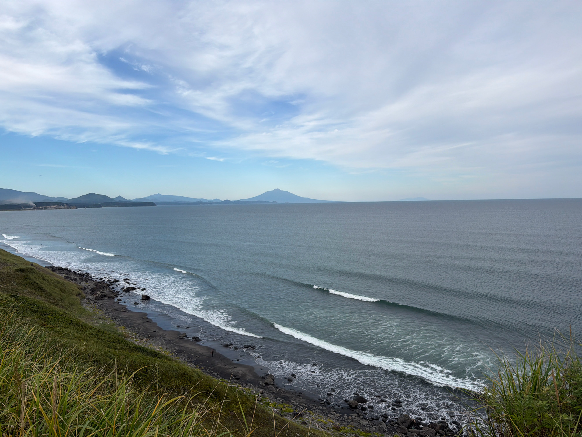 The image depicts a panoramic view of the coastline near Yuzhno-Kurilsk, Russia. A dark, rocky beach meets the turbulent ocean, where waves crash with white foam. The water is a deep, murky blue, with several lines of incoming waves visible. The coastline is covered in lush green grass and vegetation, with tall stalks of grass in the foreground partially obscuring the view. In the distance, a mountain range stretches across the horizon, with a prominent, cone-shaped mountain dominating the center of the frame. The sky above is a mixture of blue and white clouds, with wispy cirrus clouds stretching across the expanse. A single person is visible on the beach, standing near the water's edge. The overall scene conveys a sense of raw, natural beauty and rugged coastal landscape.
