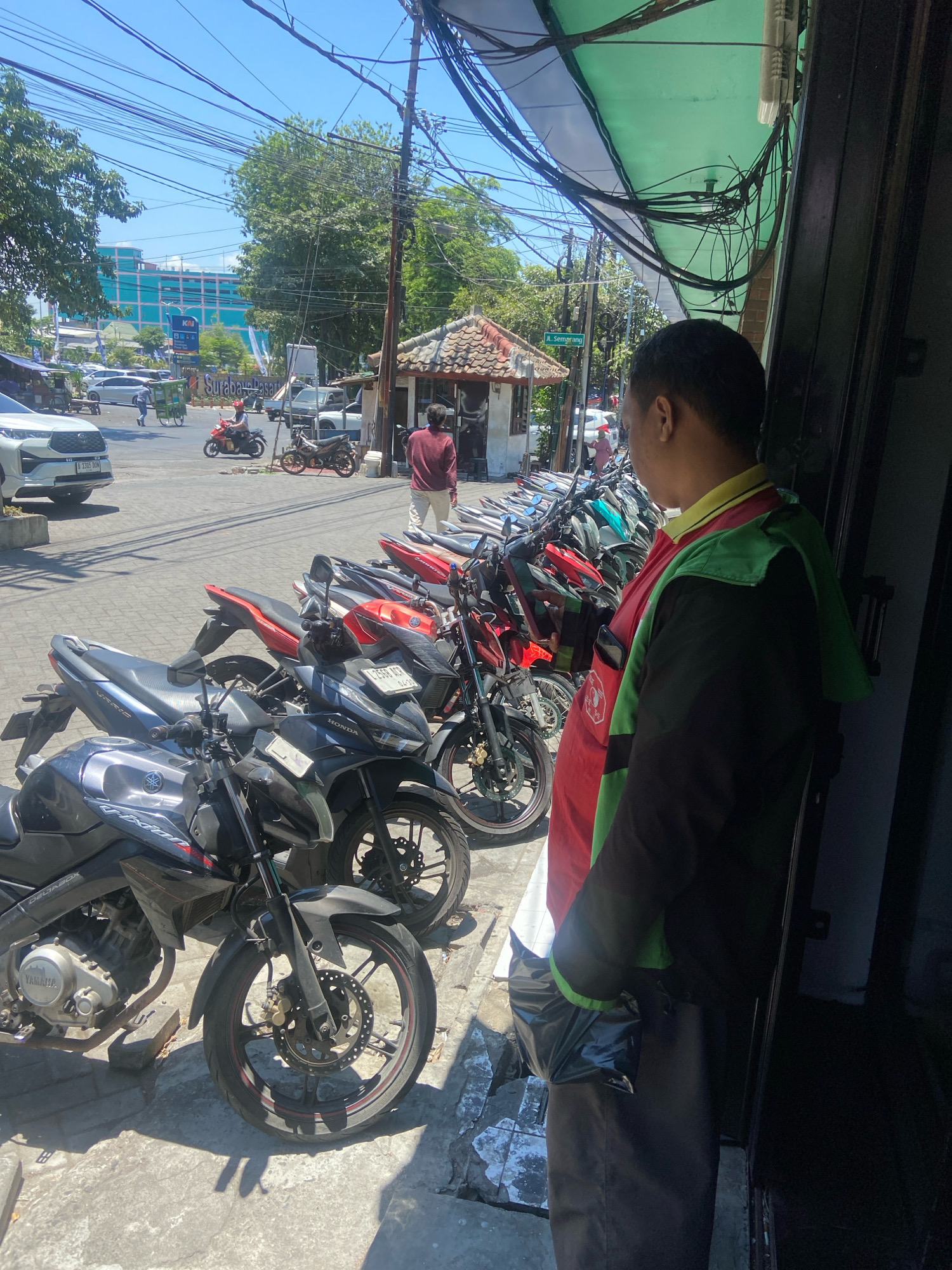 In Surabaya, Indonesia, a man wearing a bright red and green uniform with a black jacket is standing near a row of parked motorcycles. The man is facing away from the camera, and his left hand is holding a black plastic bag. The motorcycles are parked on a paved surface, and in the background, there are more motorcycles, cars, and buildings. A sign visible in the background reads "Surabaya Pasar". The scene is outdoors, under a bright blue sky with visible power lines overhead. The weather appears to be sunny and warm.