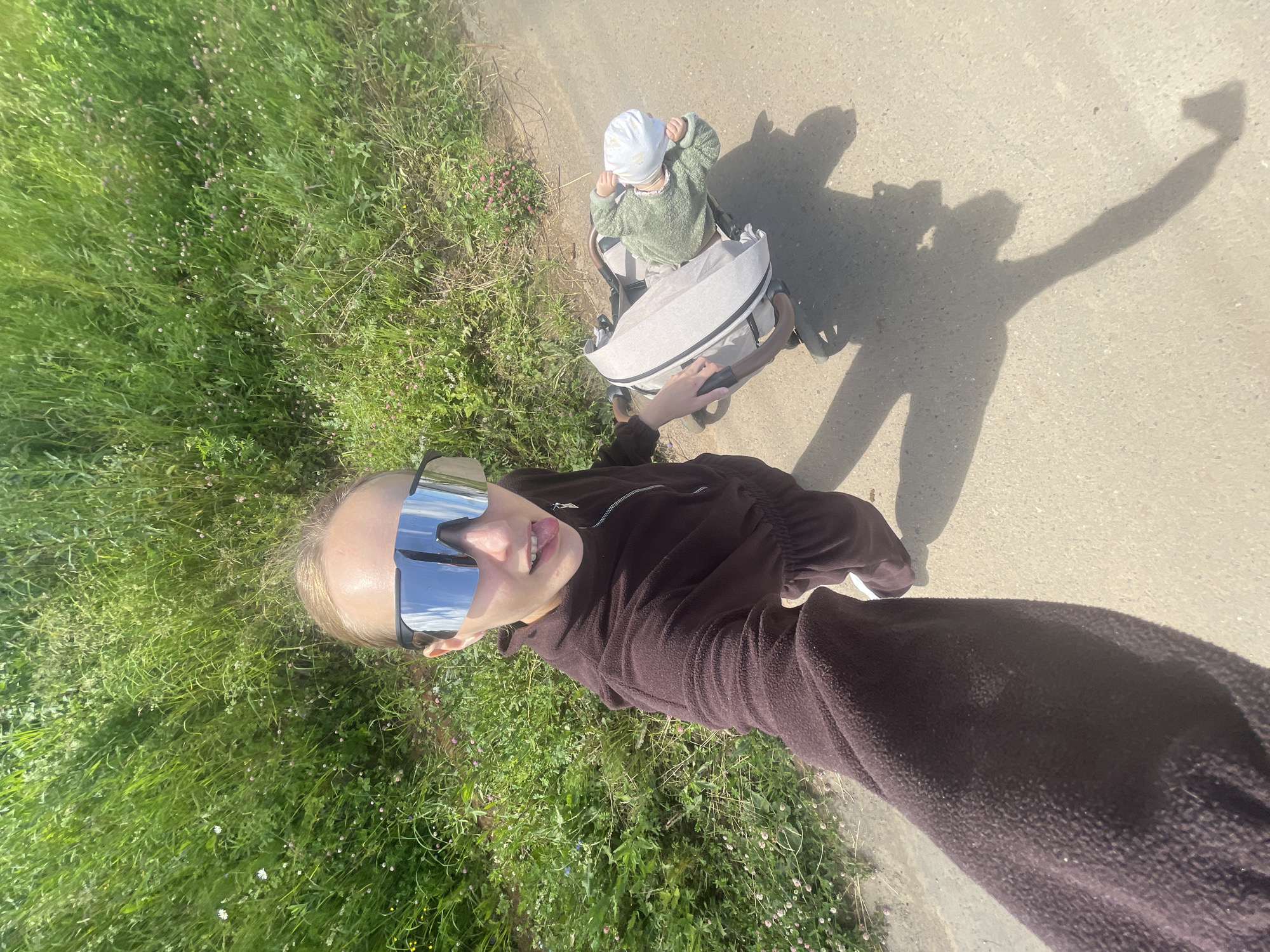 A woman and a baby are outdoors on a sunny day. The woman is in the foreground, wearing large mirrored sunglasses and a dark brown fleece jacket. She is holding a phone, taking a selfie, and has her tongue sticking out playfully. To her right, in the mid-ground, a baby sits in a stroller. The baby wears a light green sweater and a white hat. The stroller is light gray with dark brown handles. They are on a paved path next to a grassy area with wildflowers. The sun casts long shadows, indicating it is either morning or late afternoon. The scene suggests a casual outing in a park or rural area. The background is blurred green foliage and a concrete path. No text is visible. The overall mood is cheerful and relaxed.