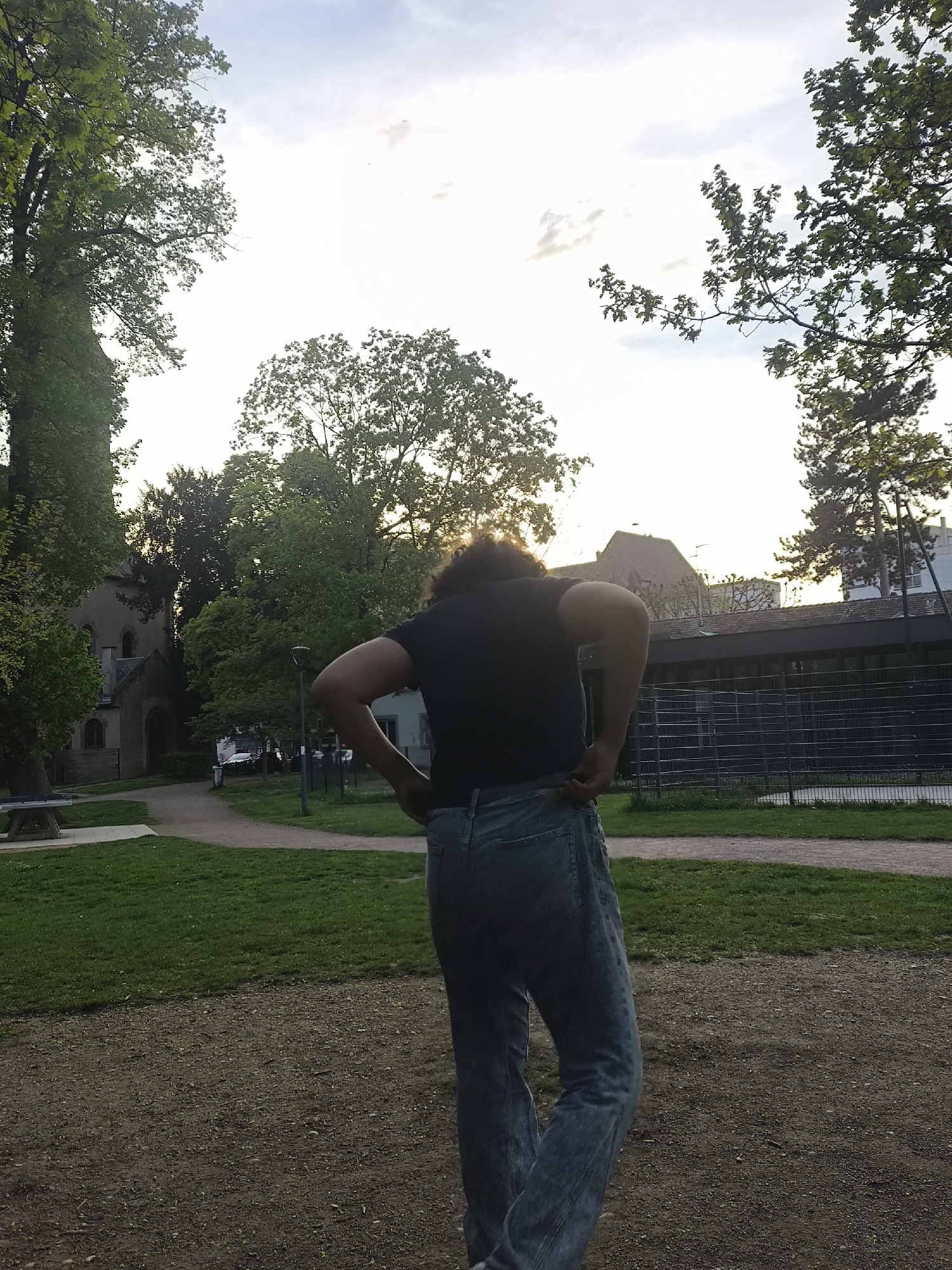 The image depicts a park scene in Strasbourg, France. A person with dark curly hair, wearing a black t-shirt and light-colored jeans, stands with their back to the camera. Their hands are on their hips. Behind the person is a green grassy area and a dirt path leading towards a building with a pointed roof, potentially a church or other religious structure. To the right, a fenced-in area with a long, low building can be seen. Trees surround the park, and the sky is bright with hints of clouds, suggesting late afternoon or early evening. A ping-pong table is visible on the left. The location is likely a public park or green space within the city of Strasbourg.