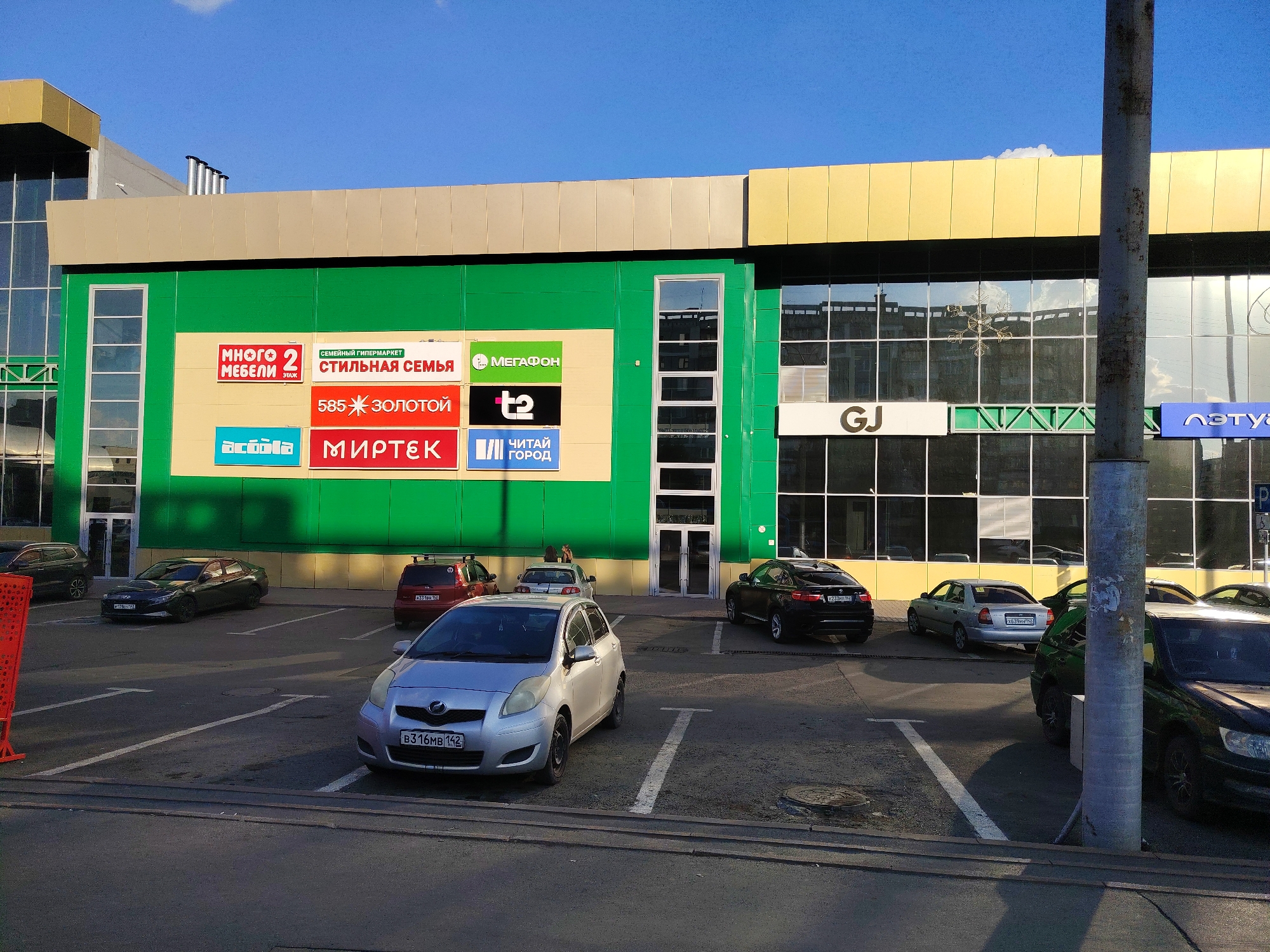 The image shows a modern shopping mall with a bright green facade, a golden trim, and large glass windows. Several cars are parked in the lot in front of the mall. The mall has multiple storefronts with various signage, including "МНОГО МЕБЕЛИ" (Many Furniture), "СТИЛЬНАЯ СЕМЬЯ" (Stylish Family), "585 ЗОЛОТОЙ" (585 Gold), "МИРТЕК" (Mirtek), "МЕГАФОН" (Megafon), "t2", and "ЧИТАЙ ГОРОД" (Read City). There is also a sign that reads "GJ" and "ЛЭТУАЛЬ" (L'Etoile). The scene is set during daylight with a clear blue sky. The parking lot is paved and marked with white lines. A lamppost stands in the foreground to the right of the image. The location is the "City Mall" in Kemerovo, Russia, situated on B. Rutger Street, 37.