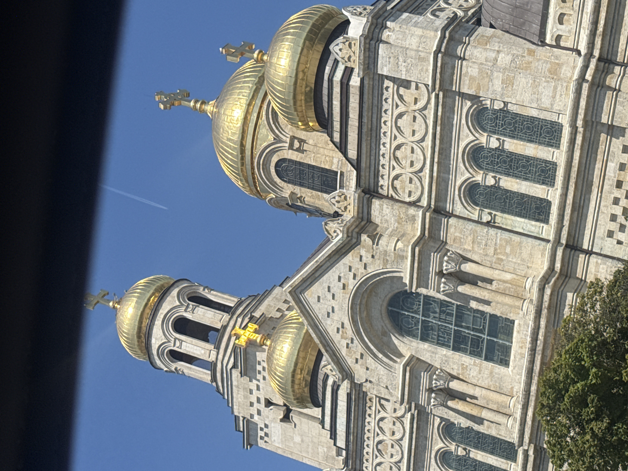 This is a close-up, angled shot of the Dormition of the Mother of God Cathedral in Varna, Bulgaria, under a clear blue sky. The image primarily features the golden domes and intricate stone architecture of the cathedral. Several golden domes, topped with crosses, are prominent. The cathedral's facade displays elaborate stonework, including arched windows with decorative patterns and pilasters. A tall bell tower, also adorned with arches and a cross, rises on the left side of the frame. A faint contrail from an airplane is visible in the upper right portion of the sky. The bottom left corner shows the top of a lush green tree. The lighting suggests it is daytime, likely late morning or afternoon, with the sun casting shadows on the stonework.