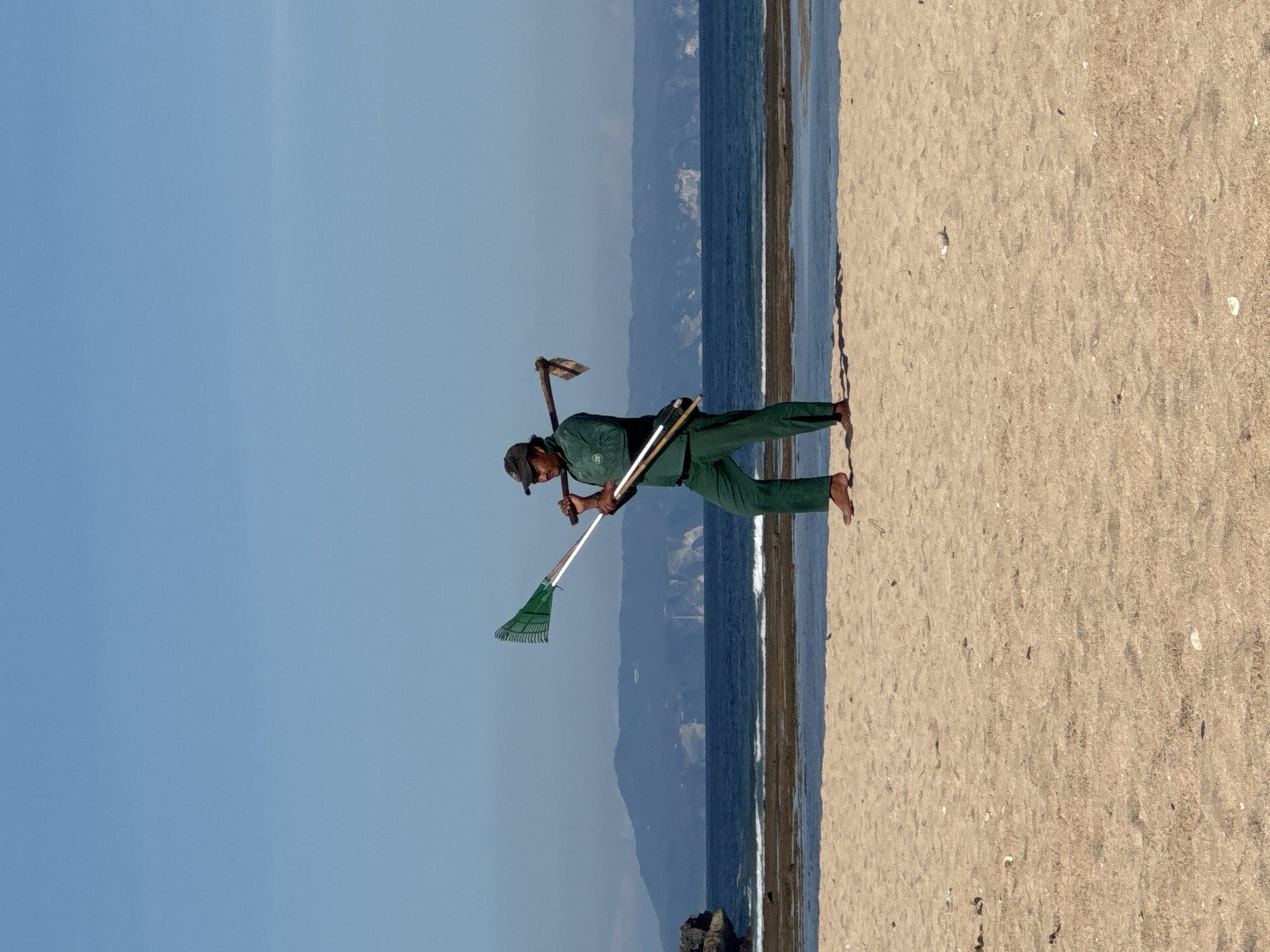 A man in green work attire stands barefoot on a sandy beach near the ocean in Kuta Selatan, Indonesia. He is carrying a rake, shovel, and what appears to be a pickaxe. The sky is blue, and the background features a mountainous landscape across the water.