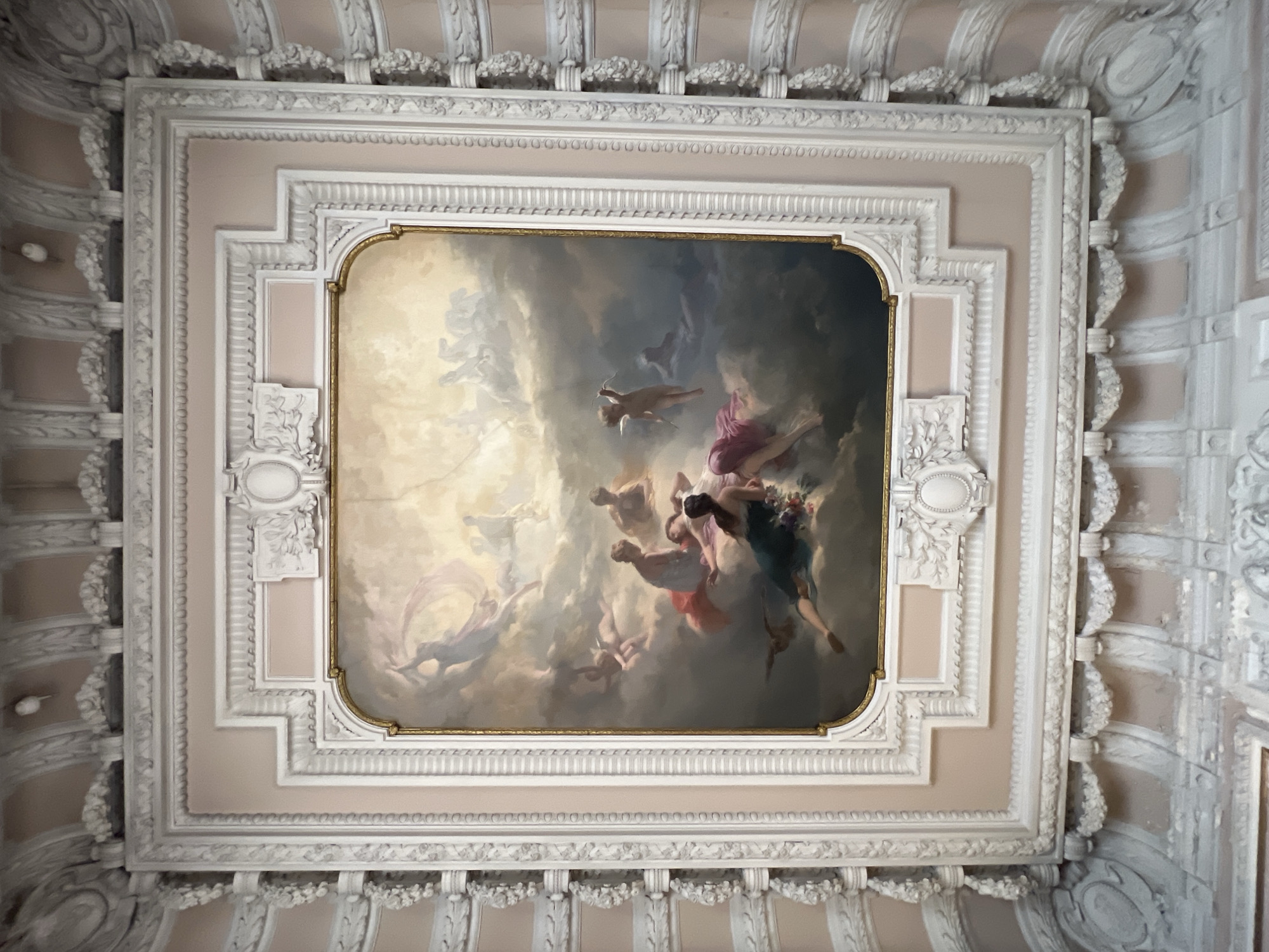 This image depicts a fresco painted on a ceiling in Saint Petersburg, Russia. The fresco features a scene with cherubic figures and women in flowing robes, rendered in a classical style with soft, ethereal lighting. The figures appear to be floating amidst clouds, suggesting a celestial or mythological theme. The fresco is set within an ornate, gilded frame and surrounded by elaborate stucco moldings that adorn the ceiling. The surrounding architecture exhibits classical influences, with intricate detailing in white plasterwork against a pale pink background. The scene is viewed from a low angle, looking upwards.