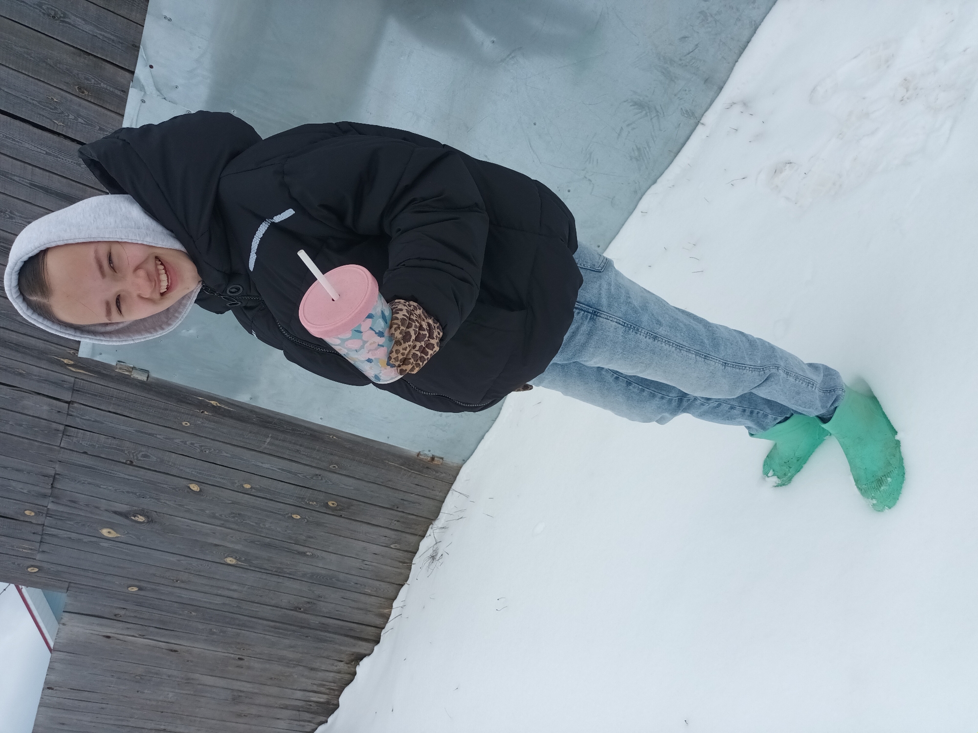 A young woman stands in the snow in Ольховка, Russia. She is wearing a black puffer jacket with a gray hooded sweatshirt underneath, light blue jeans, and light green rubber boots. She holds a pink cup with a straw and a patterned sleeve. The background includes a weathered wooden structure and a patch of gray-painted surface adjacent to the snow.