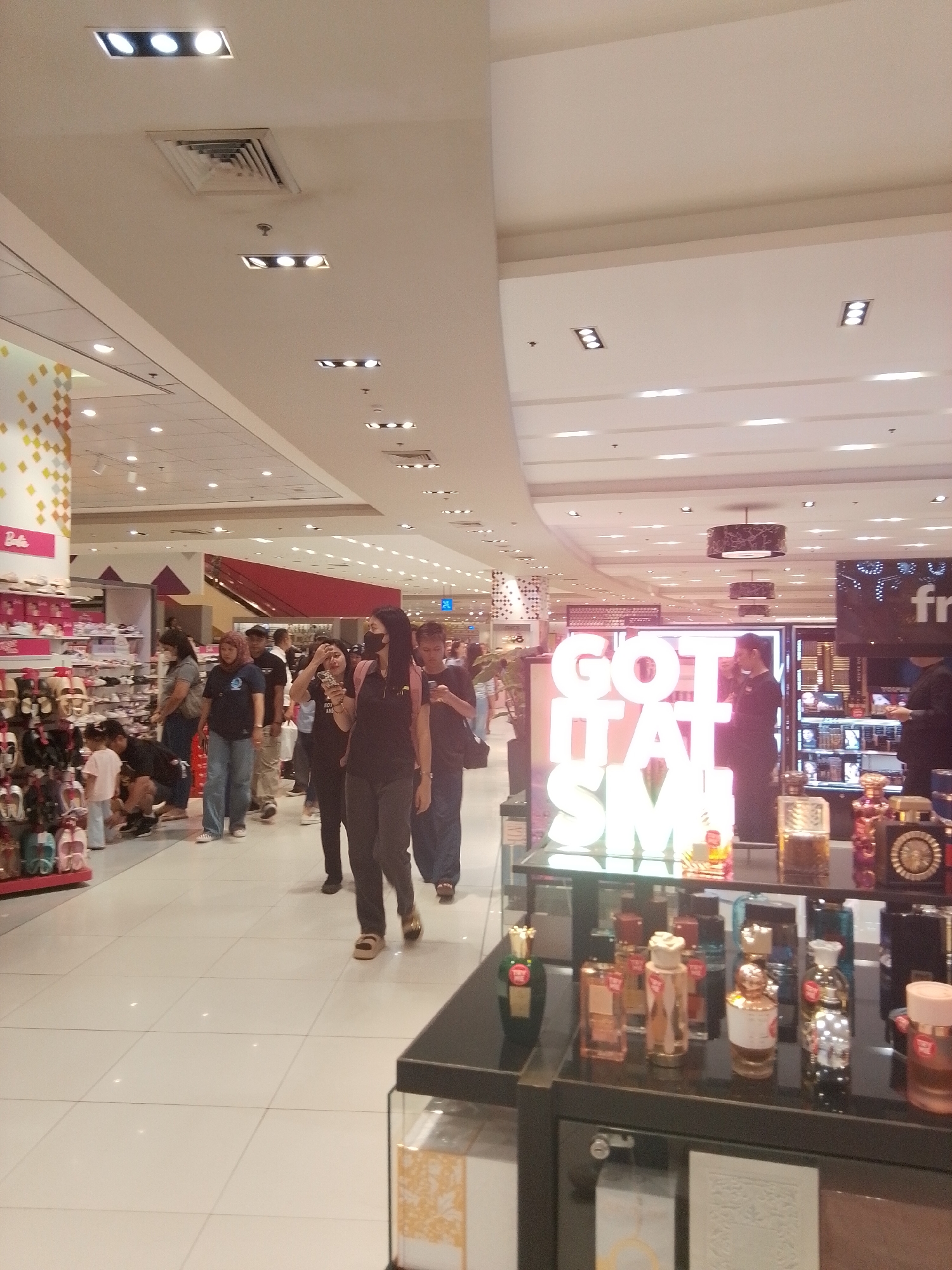 This is an indoor, eye-level photograph capturing a section of a brightly lit department store, specifically identified as an SM mall in Davao City, Philippines.
The scene is divided by a wide central aisle with a light-colored tiled floor that reflects ambient light. On the left, a shoe department is visible, featuring various footwear displayed on multi-tiered racks and shelves. A partially obscured pink sign with white lettering, reading "Barbie," is visible above a shoe display. Multiple individuals are present in this area; a child in a light pink shirt stands near a display, while an adult kneels down, seemingly examining shoes. Other adults are standing, some wearing face masks, and appear to be browsing or walking through the section.
On the right side of the aisle, a prominent, large, luminous sign with white capital letters spells out "GOT IT AT SM." In front of this sign, a black display counter features numerous perfume bottles of varying shapes and colors. Several individuals are positioned near this counter; one person stands behind it, possibly a staff member, while others are seen walking past or browsing the merchandise. Behind the "GOT IT AT SM" sign, additional retail displays with cosmetic or fragrance products are visible, along with another person looking at items.
The ceiling throughout the store is white and features an array of recessed lighting fixtures, including rectangular panels with multiple circular lights and single circular spotlights. Several air vents are also integrated into the ceiling. The overall environment is well-lit and appears to be a busy retail space.