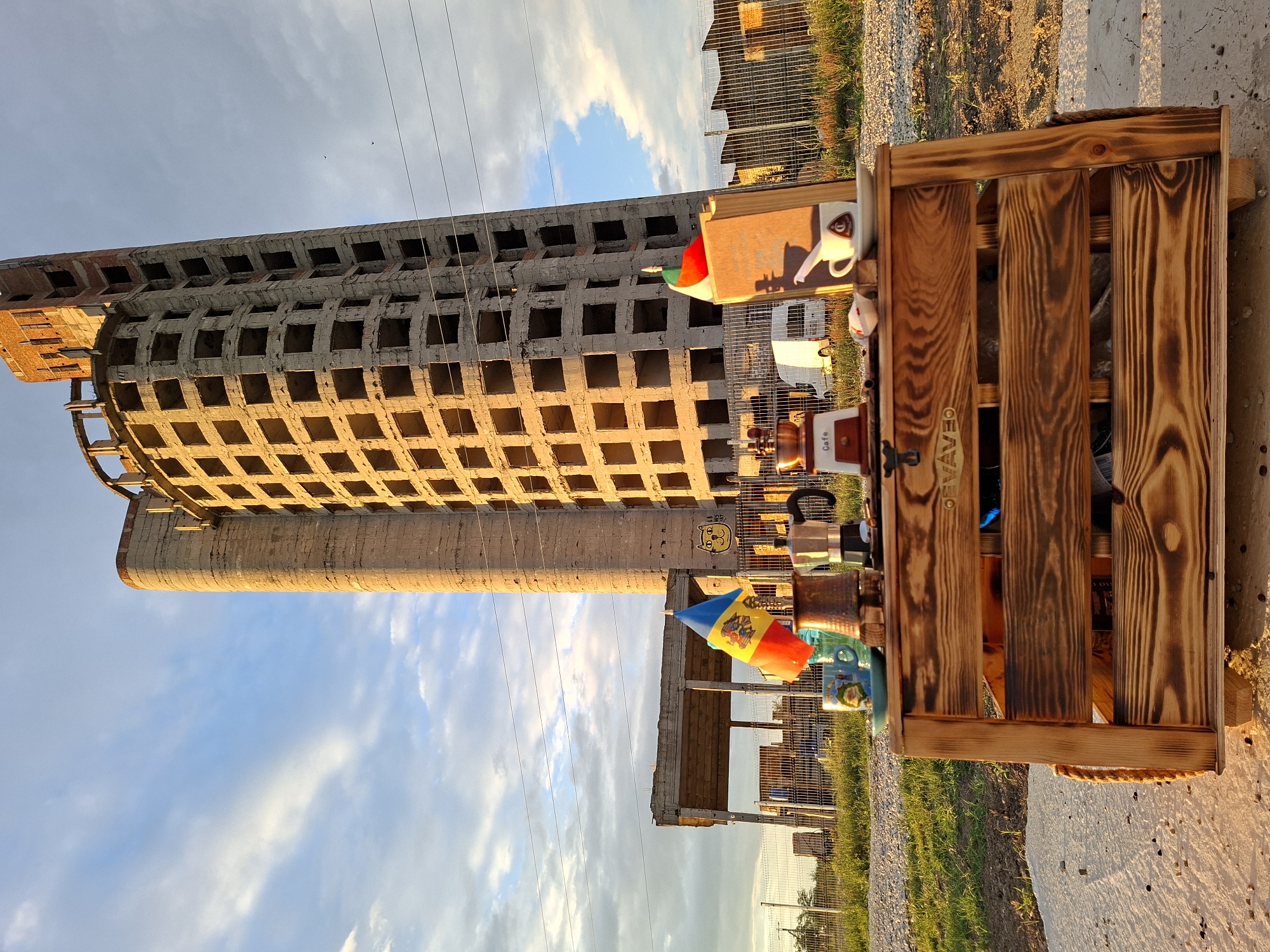 This is a wide-angle, eye-level outdoor photograph taken in Sociteni, Moldova. The scene features an unfinished, multi-story concrete building with a curved facade, appearing to be under construction or abandoned. In the foreground, a wooden cart serves as a display for coffee-related items. These include a manual coffee grinder, a moka pot, a mug with a cartoon image, a small blue saucer, a book with a flag of Moldova and another flag on top, a white cup and saucer, and a few small sugar cubes. A white van is visible in the background, behind a metal fence, and a smaller unfinished structure is also present. The sky above is partly cloudy with the sun casting a warm light, illuminating the side of the concrete building. The ground appears to be a mix of gravel and concrete.