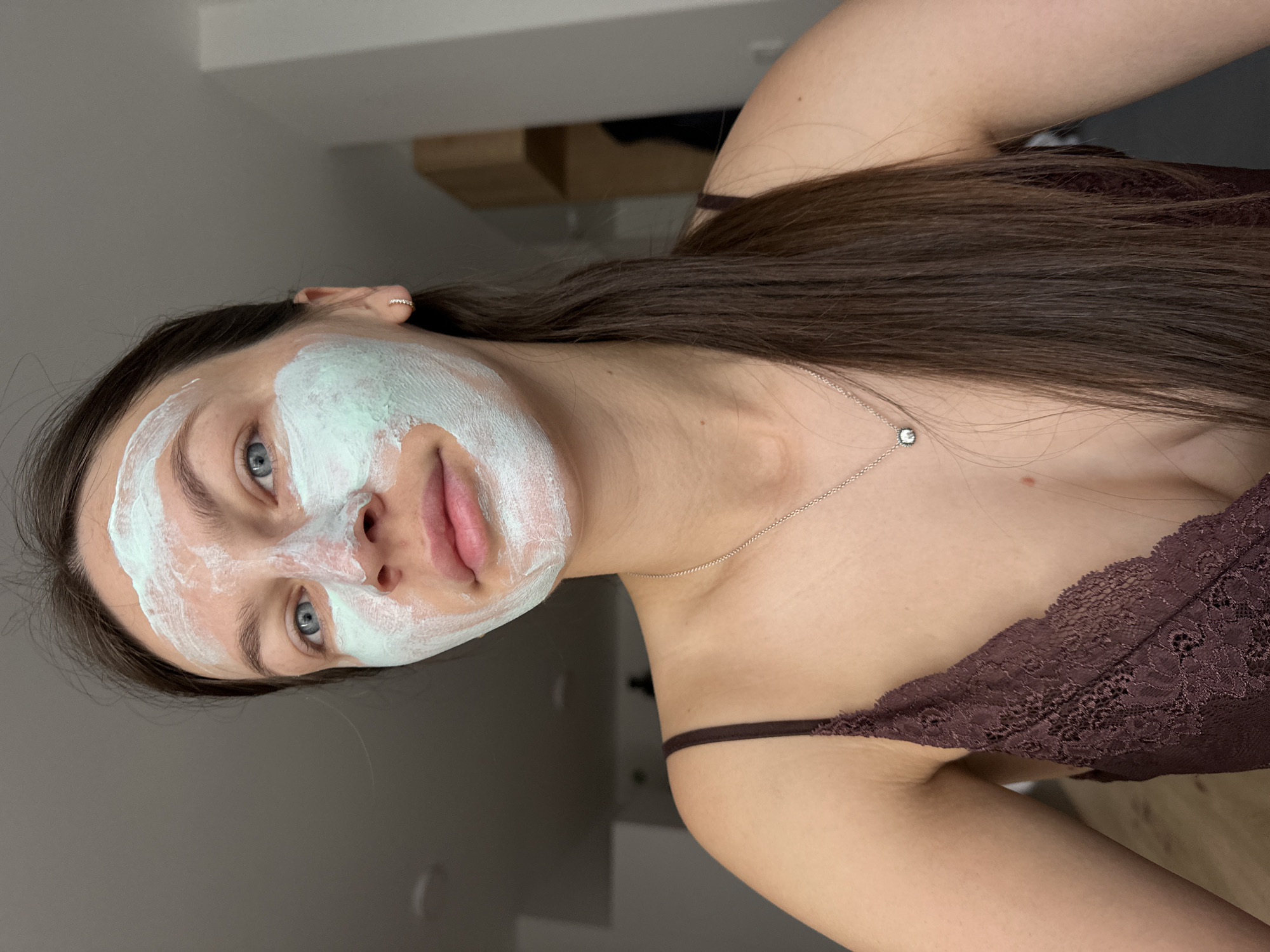 A young woman is depicted from the shoulders up, wearing a green facial mask. She has long brown hair and is wearing a brown lace top with thin straps. A delicate silver necklace with a small pendant is visible around her neck. The background is blurred, showing hints of a modern interior with gray walls and wooden furniture. There is no indication of the location being Budva, Montenegro.