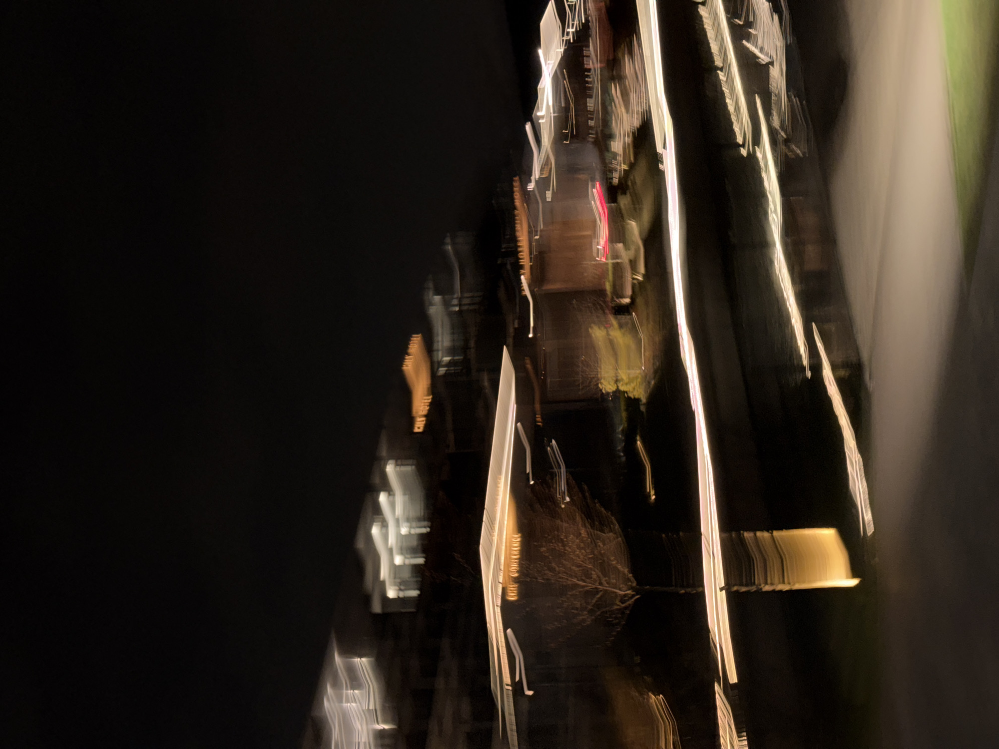 This vertical image presents an abstract night scene characterized by extreme motion blur. The dominant feature is a collection of elongated, vibrant streaks of artificial light set against a predominantly dark, black background. The left portion of the image is almost entirely black, suggesting an unlit area or sky.
The right side is filled with dynamic light trails. Predominantly white and yellow, these streaks also include hints of orange, red, and green. They appear as vertical and diagonal lines, suggesting movement either of the camera or light sources within the scene. Some of these streaks are sharp and defined, while others are broader and more diffused.
Upon closer inspection, faint outlines within the blur hint at potential subjects. In the center-right, a discernible branching pattern, illuminated by the streaking lights, strongly resembles a bare tree. Further to the left, smaller, angular white and orange light patterns might indicate distant building windows, signs, or other urban structures. Broad, soft bands of light appear on the far right, possibly originating from large light sources or illuminated surfaces.
The time of day is clearly night, evidenced by the darkness and the reliance on artificial light. There are no discernible people or specific activities taking place, with the blur rendering any potential subjects unidentifiable. No visible text is present. The image conveys a sense of speed, dynamism, and an ethereal, dreamlike quality due to the pronounced motion effect, depicting a street or urban environment at night in Vynnyky, Ukraine.