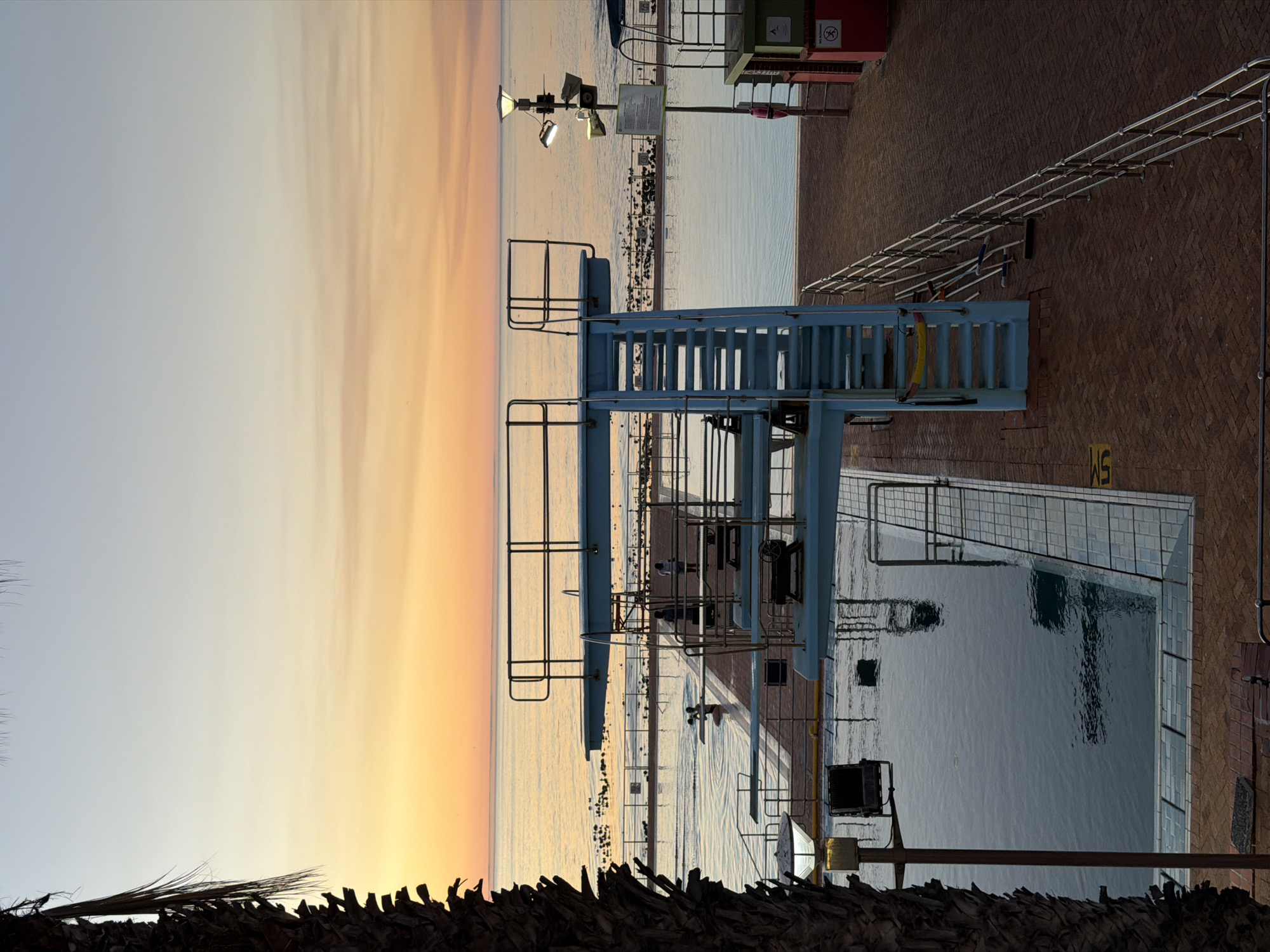 The image shows a coastal scene in Cape Town, South Africa, featuring a swimming pool complex with a diving platform. The foreground shows a brown, palm-like plant texture. To the right is a brick-paved area with metal railings. A blue diving platform stands prominently next to a swimming pool filled with water. In the background, the ocean meets a gradient sky of orange and white, indicative of sunset or sunrise. There are no visible people. The water appears still, reflecting the sky and the pool's surroundings. The picture appears to be taken from an elevated position.