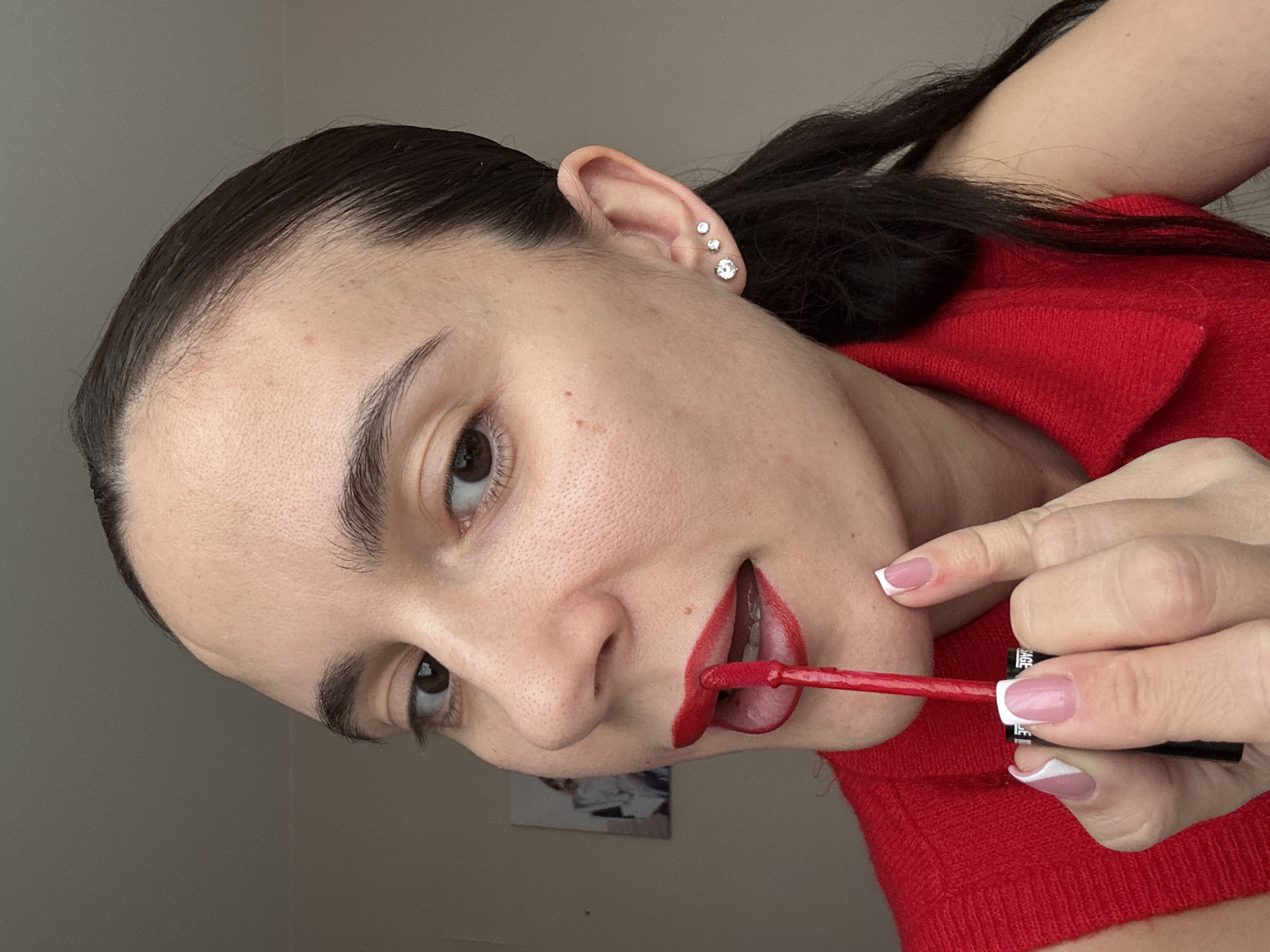 A woman is applying red lipstick. She is fair-skinned with dark brown hair pulled back. She is wearing a red sweater. She has three earrings in her left ear. Her nails are painted with a French manicure design. The background is blurry and suggests an indoor setting. There are no clear indicators of Chaikovsky, Russia in the image.
