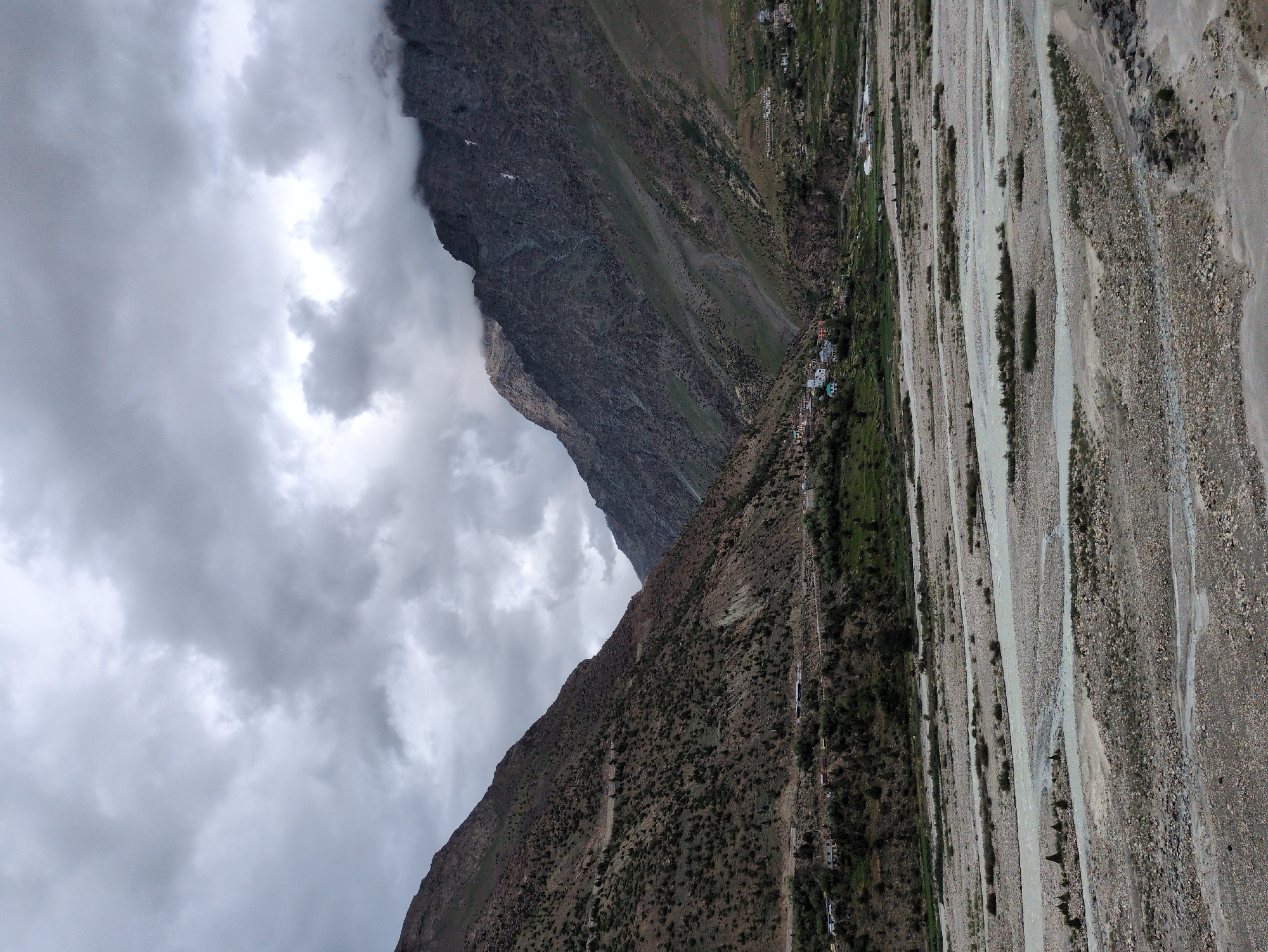 The image shows a wide, rocky riverbed in a mountainous valley. The river itself appears to be shallow and spread out, with numerous gravel bars and islands. The valley walls are steep and covered in sparse vegetation, with some evidence of human settlement on the lower slopes. In the foreground, the riverbed is dry and rocky. In the midground, the river flows in a braided channel. In the background, the valley narrows and the mountains rise steeply on either side, with a village visible on the left bank. The sky is overcast with heavy clouds, suggesting a moody or potentially rainy day. This scene is consistent with the landscape of Darcha, India, a village located in the Lahaul and Spiti district of Himachal Pradesh, known for its high-altitude valleys and glacial rivers.