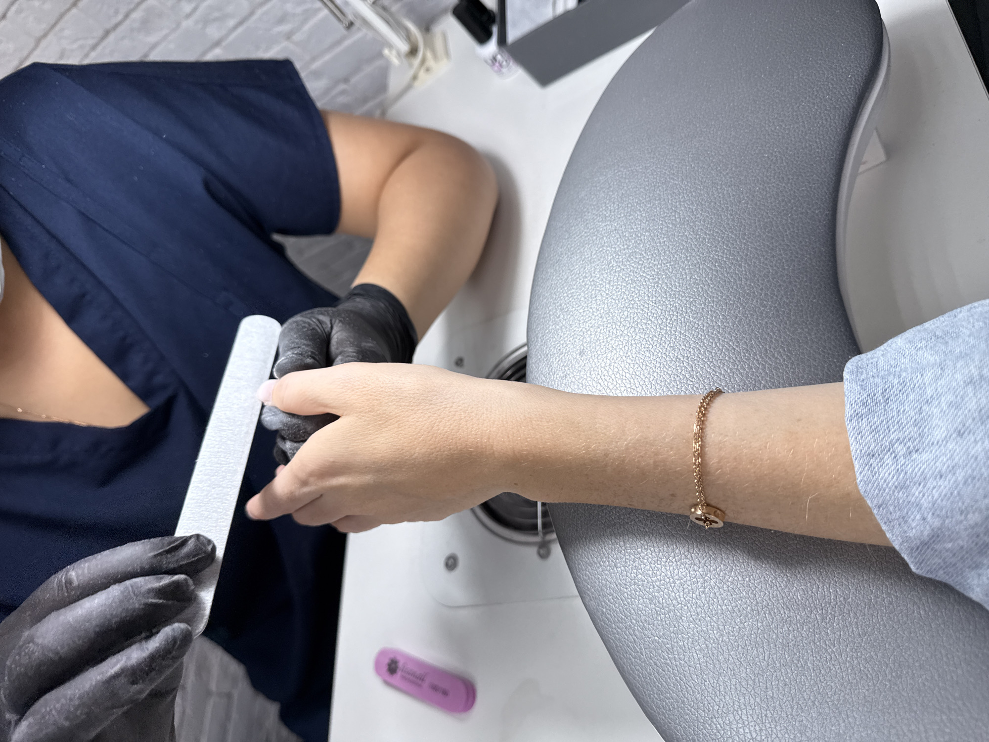 An overhead view captures a professional manicure session in what appears to be a nail salon or beauty studio in Moscow, Russia. The scene focuses on two individuals: a nail technician and a client.
The nail technician, positioned on the left side of the frame, is wearing a dark blue scrub top and black protective gloves. Both gloved hands are actively engaged in shaping the client's nails with a long, white, fine-grit nail file. The technician's posture suggests focused attention on the task.
The client's right arm extends into the frame from the right, resting on a textured, grey arm support that is part of a white manicure table or workstation. The client's arm is partially covered by a light blue sleeve, and a delicate golden chain bracelet with a small, circular charm (possibly a star or compass design) adorns their wrist.
In the background, a white wall with a subtle brick-like texture is visible. On the white manicure table, a small pink nail buffer or file is placed, with the text "Formula pro" and "100/180" faintly legible, along with a floral or star-like logo. The lighting is bright and even, indicative of an indoor setting, likely during the day. The overall impression is one of a professional and clean environment where beauty services are being performed.
