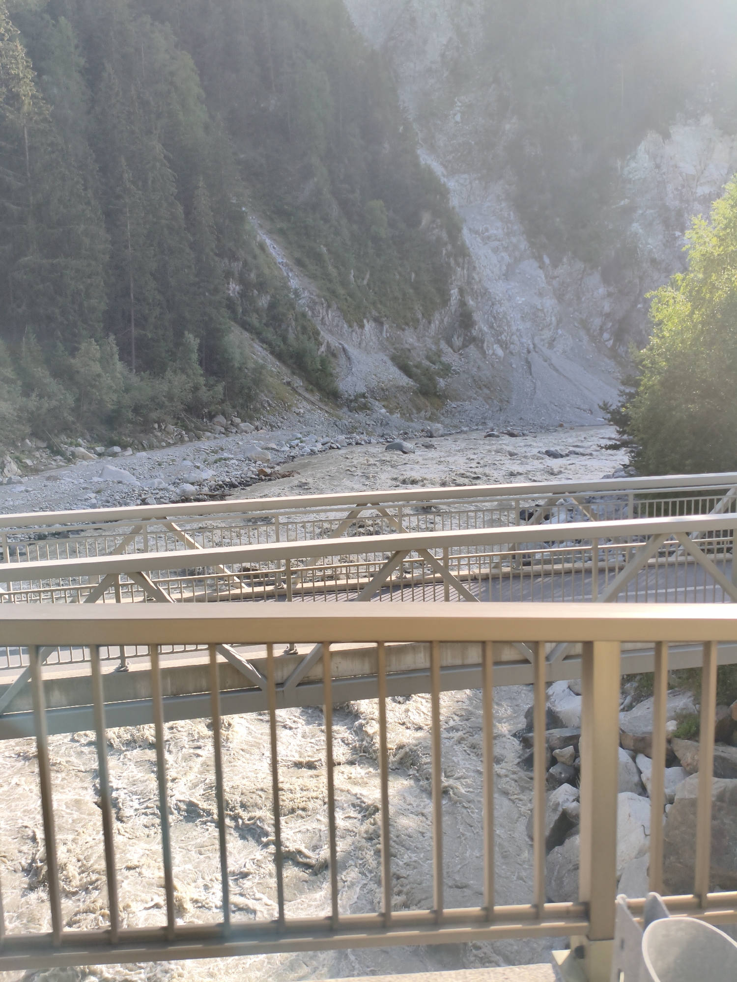 The image shows a wide, fast-flowing river with muddy, turbulent water, running through a gorge. Steep, rocky hillsides covered in dense green trees rise on either side of the river. Two parallel metal bridges span the river, with the foreground bridge clearly visible, featuring a light-colored railing. The sun is shining, creating a bright haze in the upper right portion of the image. The scene appears to be a natural landscape with man-made infrastructure for crossing. This location is the Winkl bridge over the Drava river in Winklen, Austria.