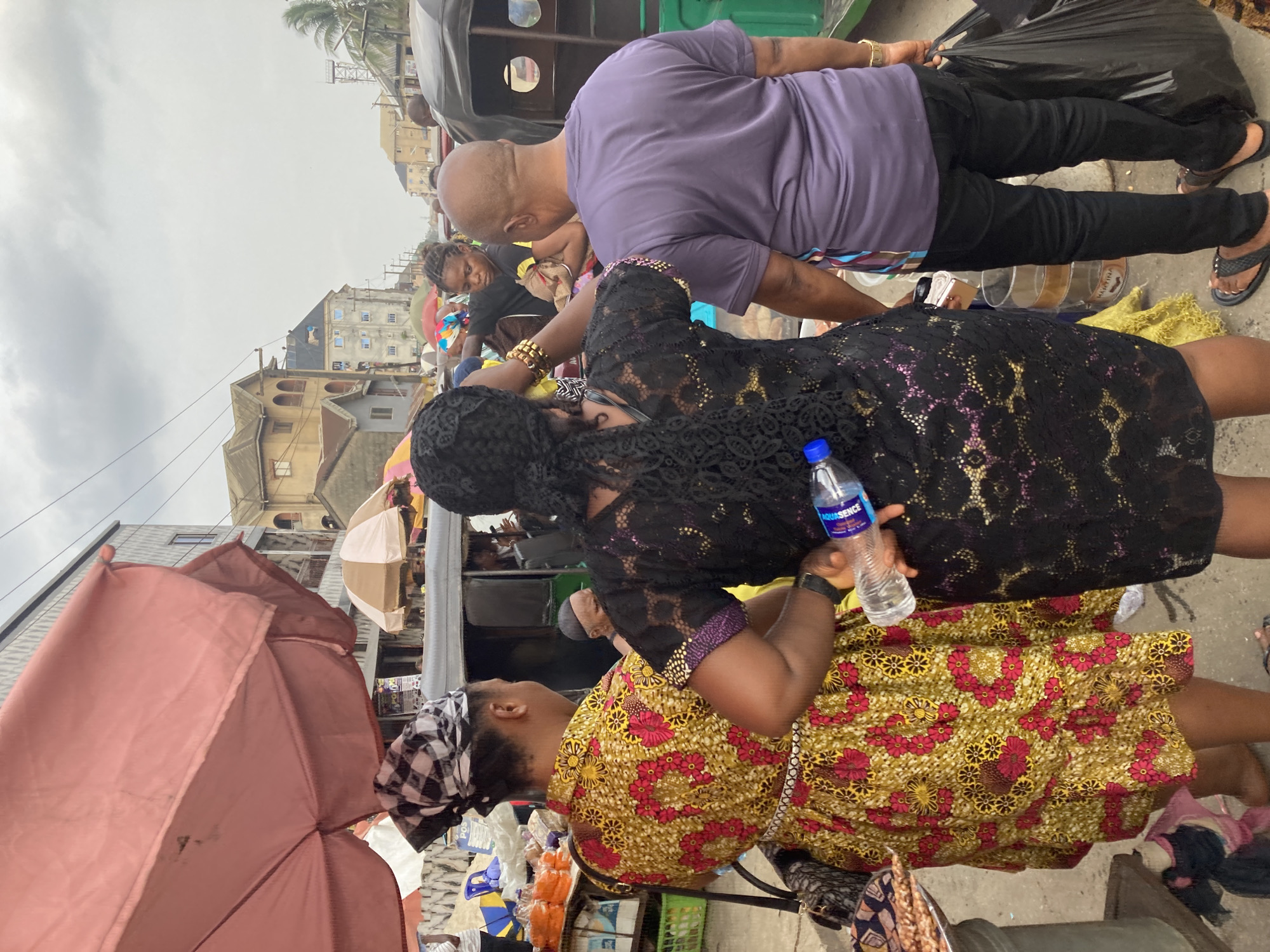 A busy street scene in Ura Nta Umuarandu, Nigeria. A woman in a black lace dress holds a bottle of "Aquasence" water, standing next to another woman wearing a yellow and red floral dress with a black and white headscarf. A man in a purple shirt and black pants stands nearby, holding a black plastic bag. The scene is crowded, with umbrellas providing shade and buildings visible in the background.