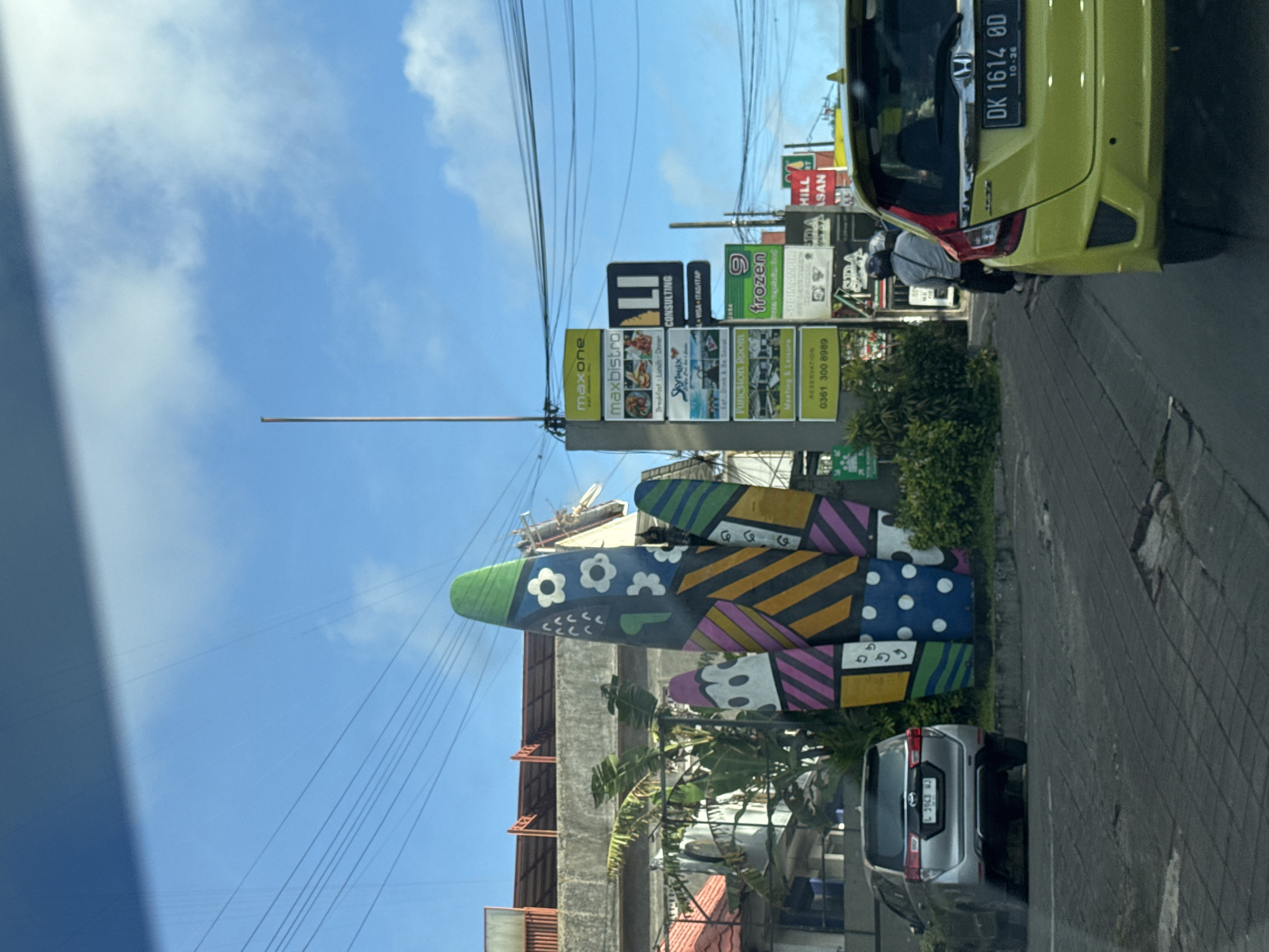 A street scene in Kuta Selatan, Indonesia, shows an intersection with signage for various businesses, including Maxone, Maxbistro, Skymax, Frozen, and others. A large, decorative sculpture of fish with colorful geometric and floral patterns stands prominently near the intersection. Utility poles and wires are visible overhead. Vehicles, including a yellow car and a silver car, are positioned along the street. The sky is blue with scattered clouds.