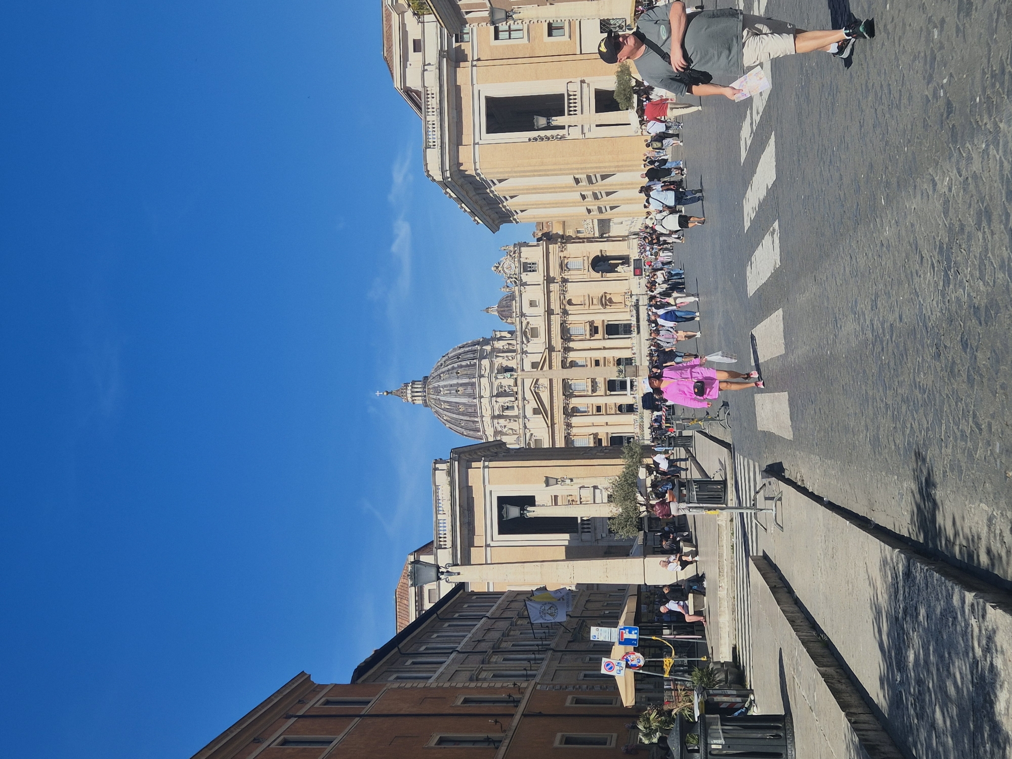 The image shows St. Peter's Basilica in Vatican City, Rome, from across the street. The sky is clear blue, and the buildings are made of light-colored stone. A crowd of people is gathered in the street, and a person wearing a pink outfit is walking on the crosswalk. In the foreground, a street lamp, road signs, and other city furniture can be seen along with the shadow cast by a tree. A man wearing shorts and a gray shirt is standing to the right, looking at the basilica.