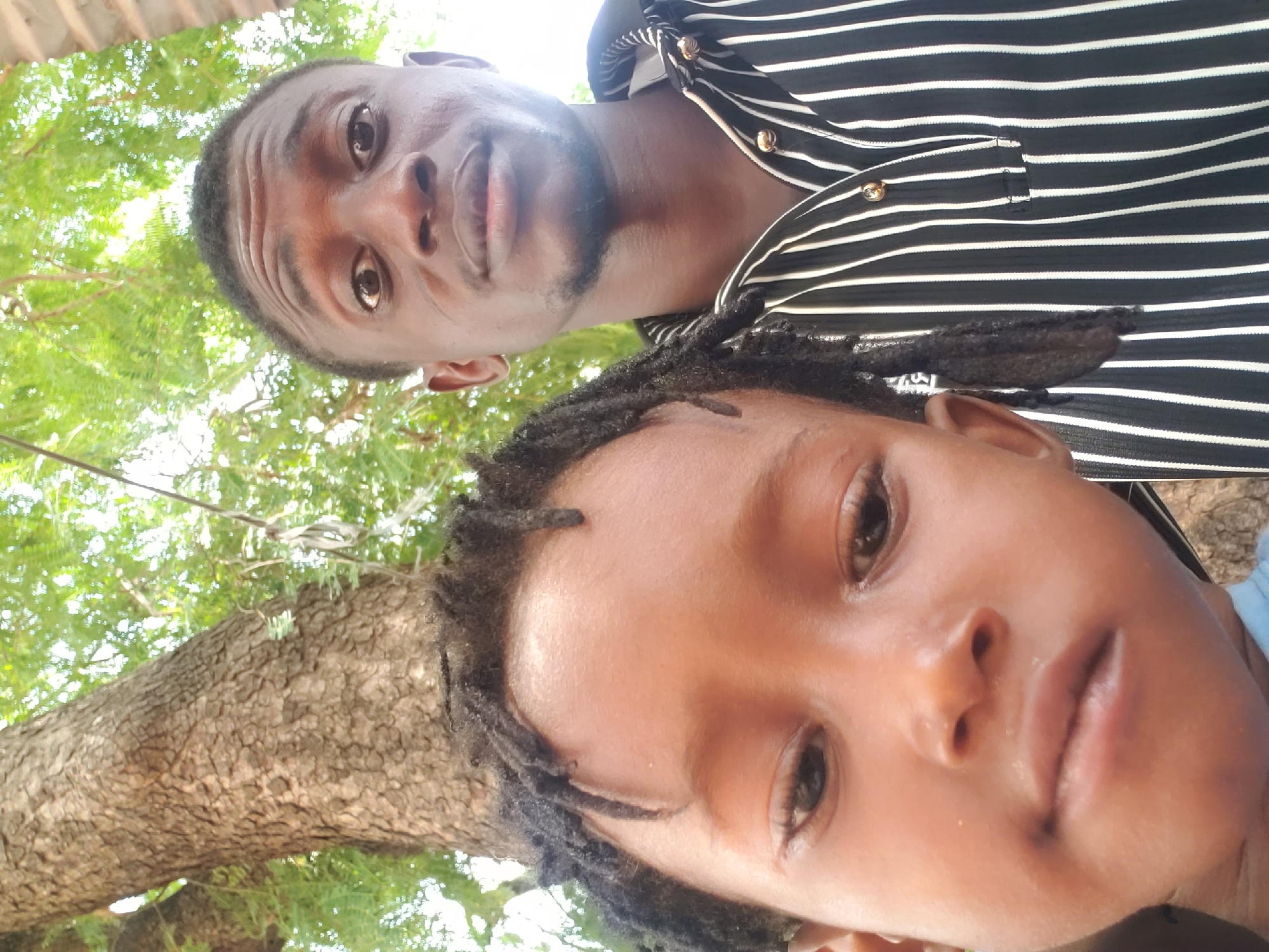 The image presents a distinctive low-angle portrait of two individuals, an adult male and a younger person, captured outdoors in Adankolo, Nigeria. Both subjects are looking directly into the camera, suggesting a selfie taken from a ground-level or very low perspective.
The adult male, positioned towards the top of the frame, has dark skin, short dark hair, and a trimmed beard or goatee. He wears a black shirt with horizontal white stripes and prominent gold-colored buttons on the placket. His expression is neutral and direct.
Below him, closer to the lens, is a younger individual with dark skin and dark hair styled in dreadlocks. Their face, with open eyes and slightly parted lips, fills a significant portion of the lower frame. A fragment of a light blue garment is visible near their shoulder.
The background is natural, dominated by vibrant green tree leaves and the textured bark of a tree trunk, indicating the photograph was taken beneath a tree canopy. The bright, even lighting suggests it is daytime with clear weather. No specific activity beyond posing for the photograph is evident, and there is no discernible text within the image.