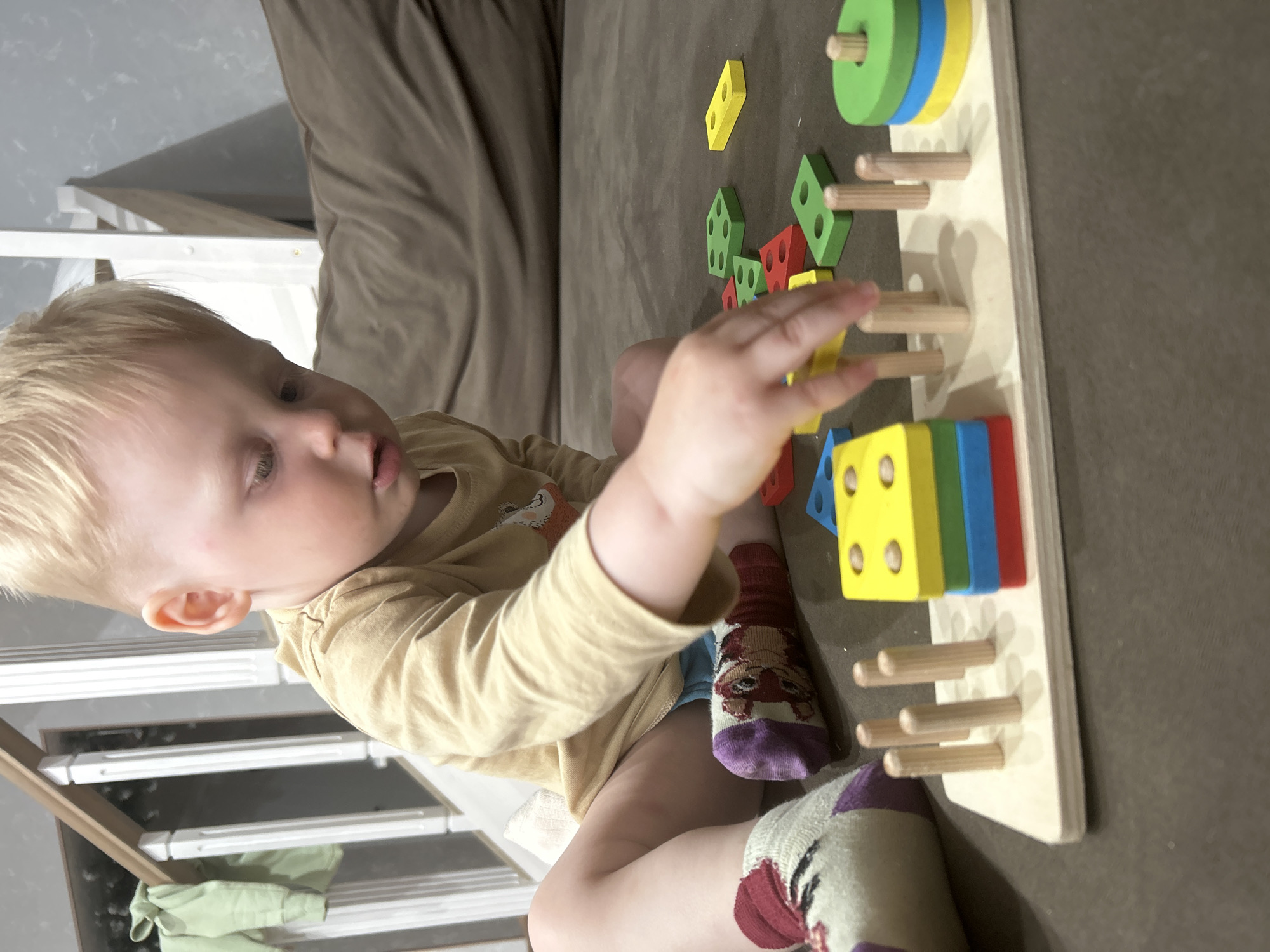 A young child with blonde hair, wearing a tan shirt and socks with animal designs, is seated on a brown sofa. The child is interacting with a wooden toy consisting of a rectangular board with pegs, and colorful geometric shapes. The setting appears to be indoors. Location: unknown.