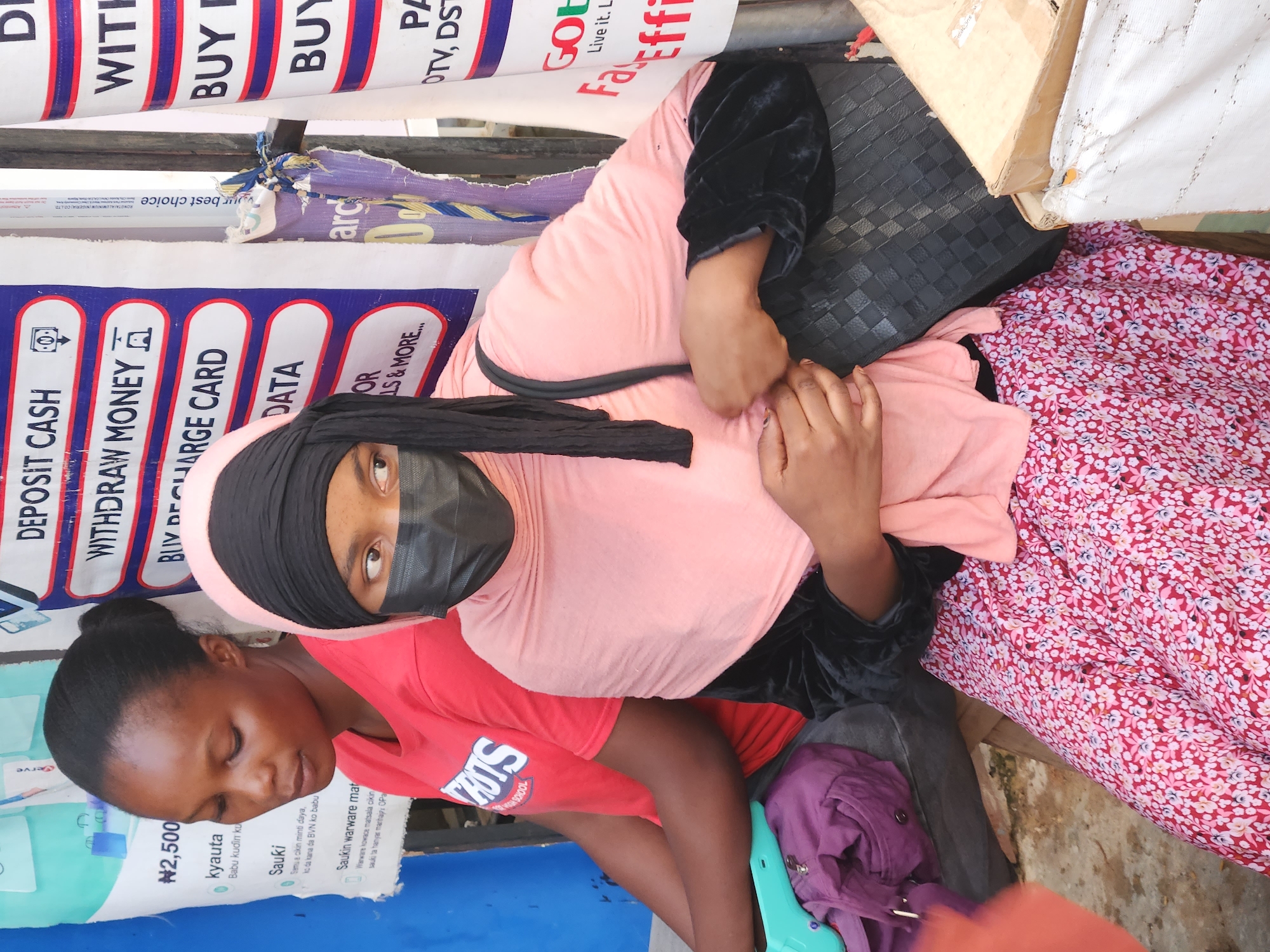 Two women are seated next to each other outdoors in an urban environment. One woman is wearing a pink hijab, a black face mask, and a black velvet top. She is sitting next to a black purse and a floral print dress. The other woman is wearing a red T-shirt with "SLIS" printed on it. A sign behind them advertises services such as "DEPOSIT CASH," "WITHDRAW MONEY," and "BUY RECHARGE CARD." Additional signs advertise "GOtv" and "FacEffi." Some items are on the ground, including a purple jacket and a turquoise object.