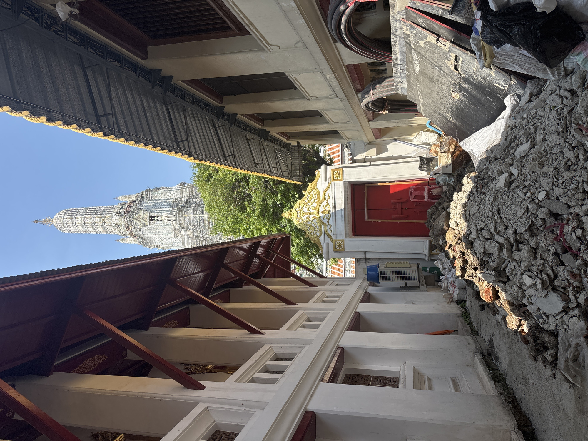 The image depicts a view of Wat Arun Ratchawararam Ratchawaramahawihan, a Buddhist temple in Bangkok, Thailand, seen from an alleyway between buildings. The temple's central prang (tower) is visible against a clear blue sky, partially obscured by a green tree.
The alleyway is cluttered with construction debris, including broken bricks and concrete rubble, and trash bags. The buildings on either side of the alleyway appear to be under construction or renovation. One building has a red door and a white facade. The other features a metal awning and a decorated roof.
