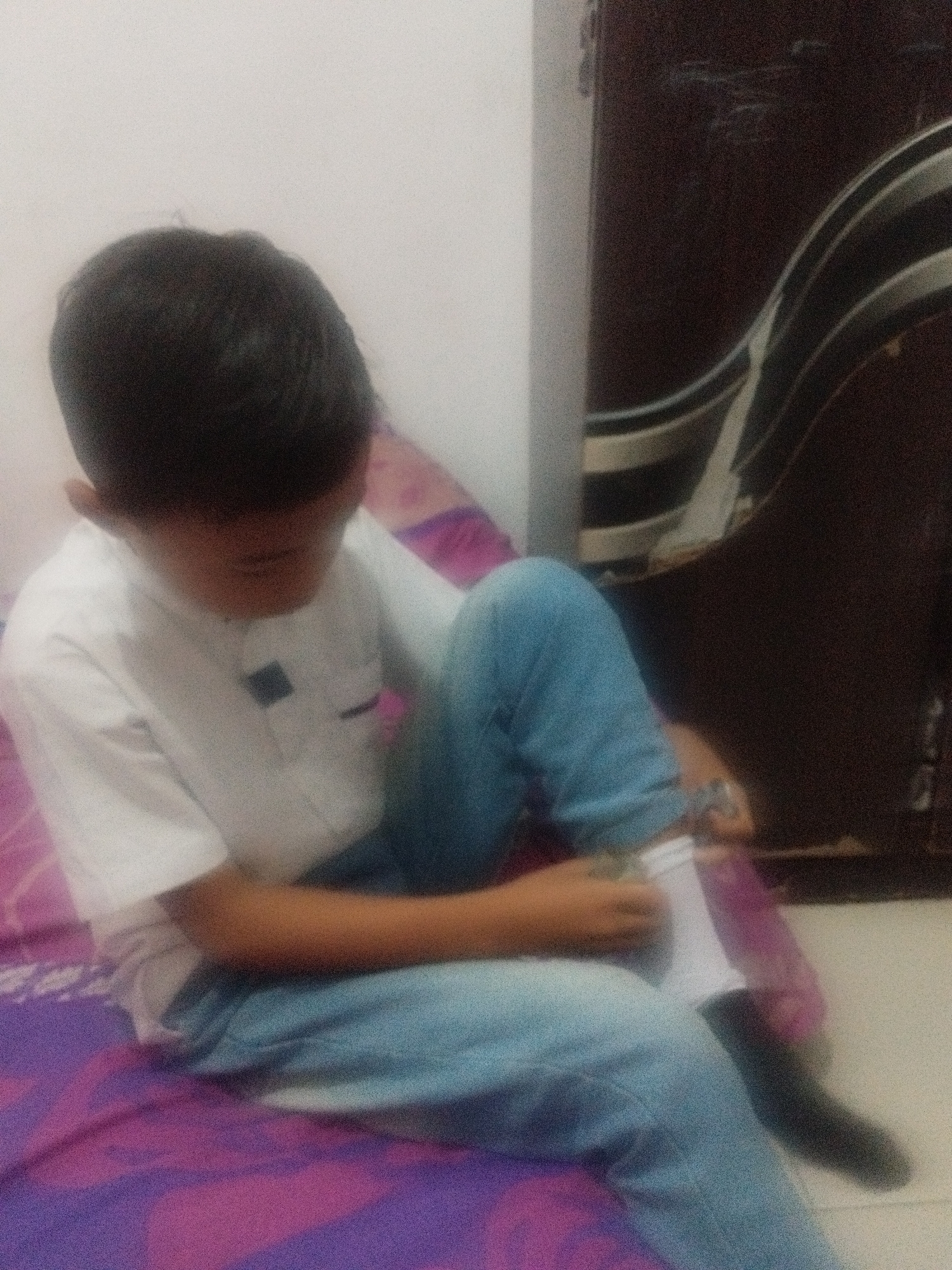 In Surabaya, Indonesia, a young boy is sitting on a bed with a purple and pink patterned sheet. He is wearing a white shirt and blue jeans. His left leg is bent, and he is holding a white object in his hands, appearing to be putting on a sock. The background is dimly lit, with a dark, wooden cabinet visible to the right. The scene suggests a casual, indoor setting, possibly a bedroom.