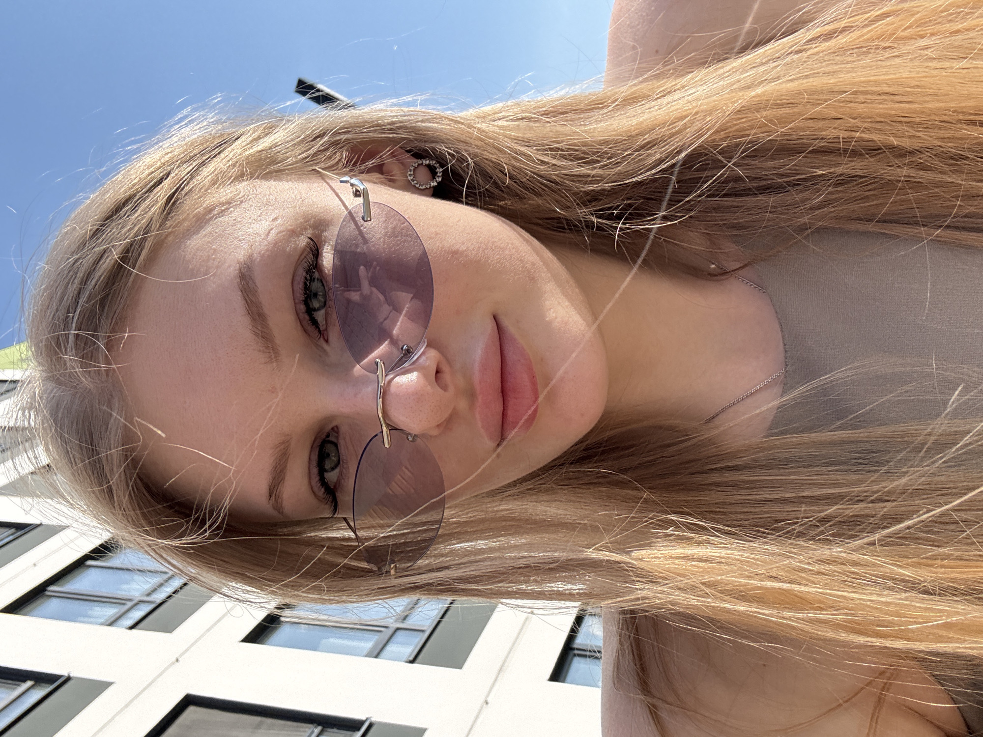 A young woman with long blonde hair and round sunglasses is angled in an outdoor shot. Her face fills the majority of the frame. She wears a light-colored top and a silver necklace. Behind her is a multi-story building with windows, and above, a clear blue sky. The scene is likely in daylight. The city and specific location within Perm, Russia cannot be determined from the visual information provided.