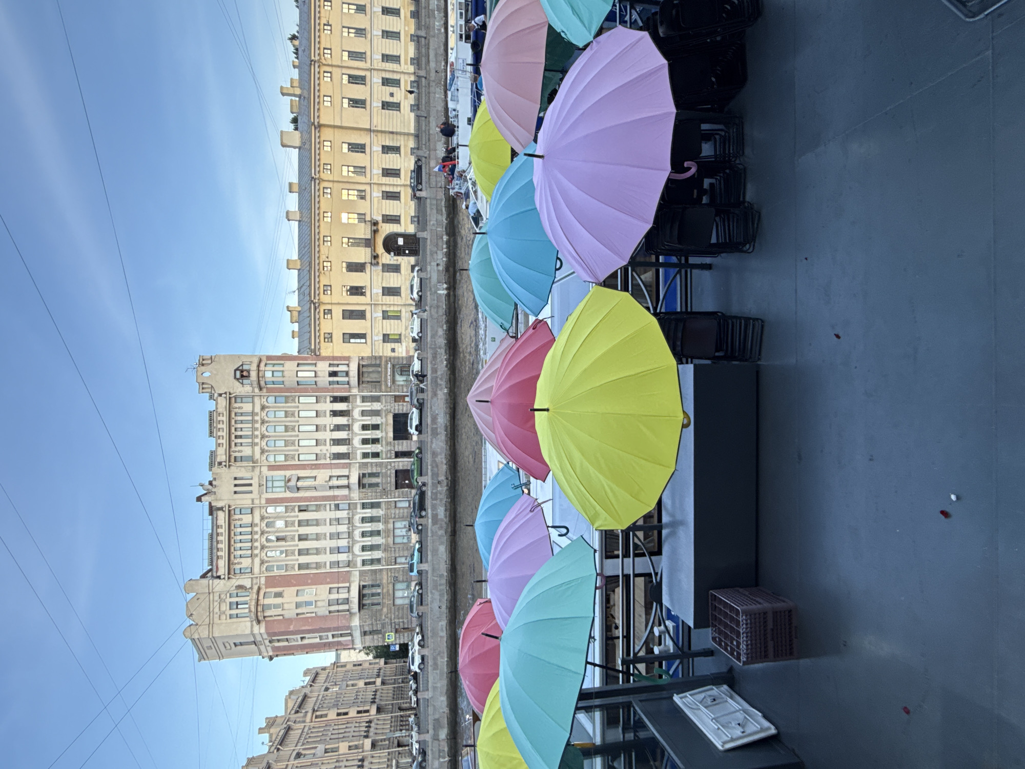 The image depicts a view from a vessel on the Neva River in Saint Petersburg, Russia, looking towards the embankment. In the foreground, a deck is adorned with a vibrant assortment of open umbrellas in various pastel shades, including yellow, turquoise, pink, and lavender. Stacks of folded black chairs are visible beneath some of the umbrellas, suggesting preparations for an event or seating for passengers. A dark gray rectangular container and a white foldable board are also present on the deck. Beyond the umbrellas, the Neva River flows, with several boats and a few people visible on them. The embankment features a pedestrian walkway lined with cars. In the background, imposing historic buildings with ornate facades and numerous windows stand tall against a clear blue sky, with overhead wires crisscrossing the frame. The architecture suggests the presence of classical and Art Nouveau styles, characteristic of Saint Petersburg. The overall scene captures a lively and colorful atmosphere on the river, with the grand architectural heritage of the city serving as a backdrop.