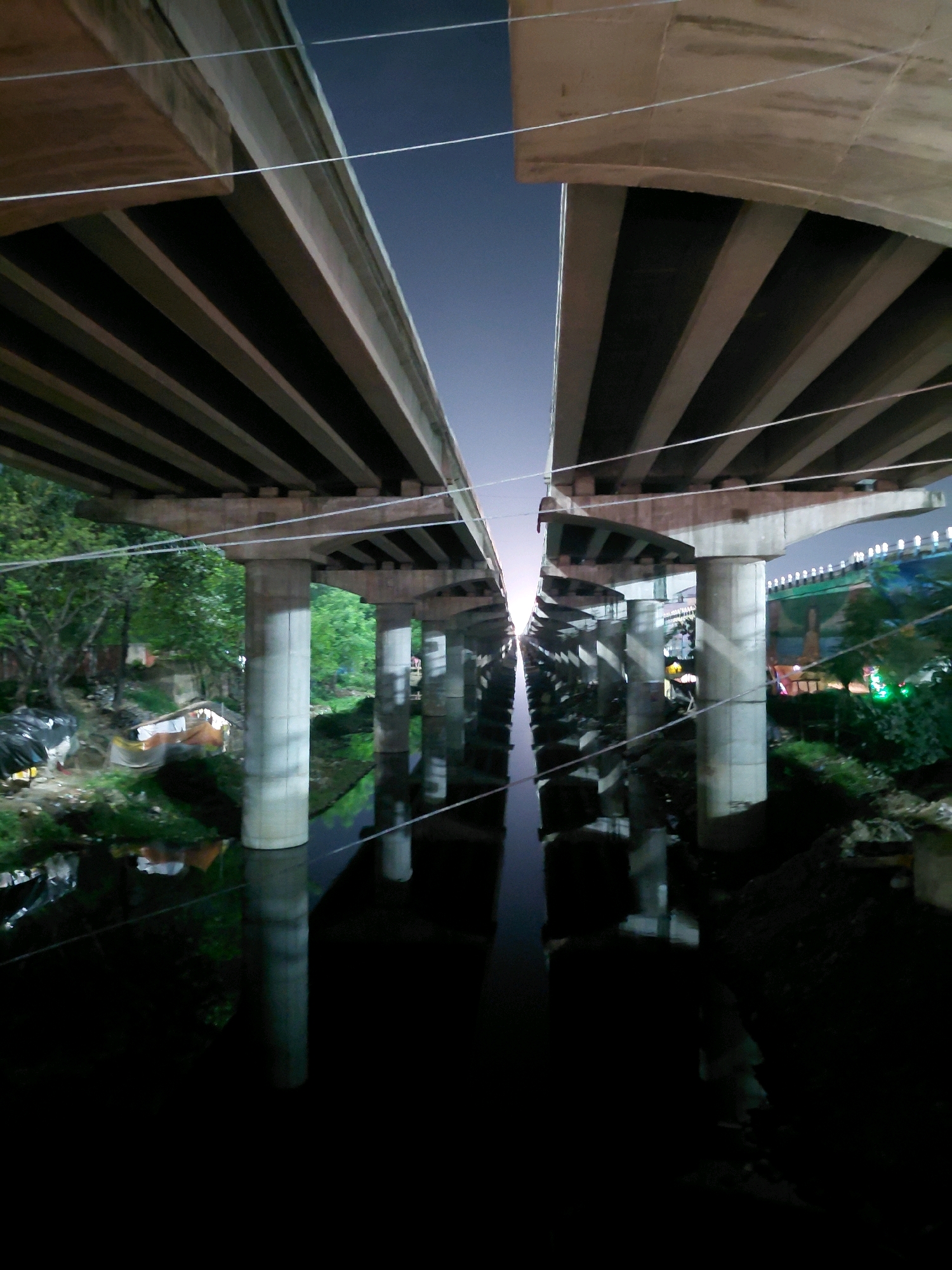 This is a nighttime photograph taken from below an elevated highway in Patna, India, likely along the Ganga River. The highway is constructed of concrete with visible supporting pillars and beams. The sky above is dark and clear. Below the highway, a body of water, likely a river or canal, reflects the overhead structure and the surrounding environment. On the banks of the water, there are signs of informal settlements with makeshift structures and debris, indicating a densely populated urban area. Lush green trees are visible on one side of the riverbank, contrasting with the concrete infrastructure. In the distance, lights and some structures suggest further urban development. The perspective is low and wide, emphasizing the scale of the highway and its shadow.